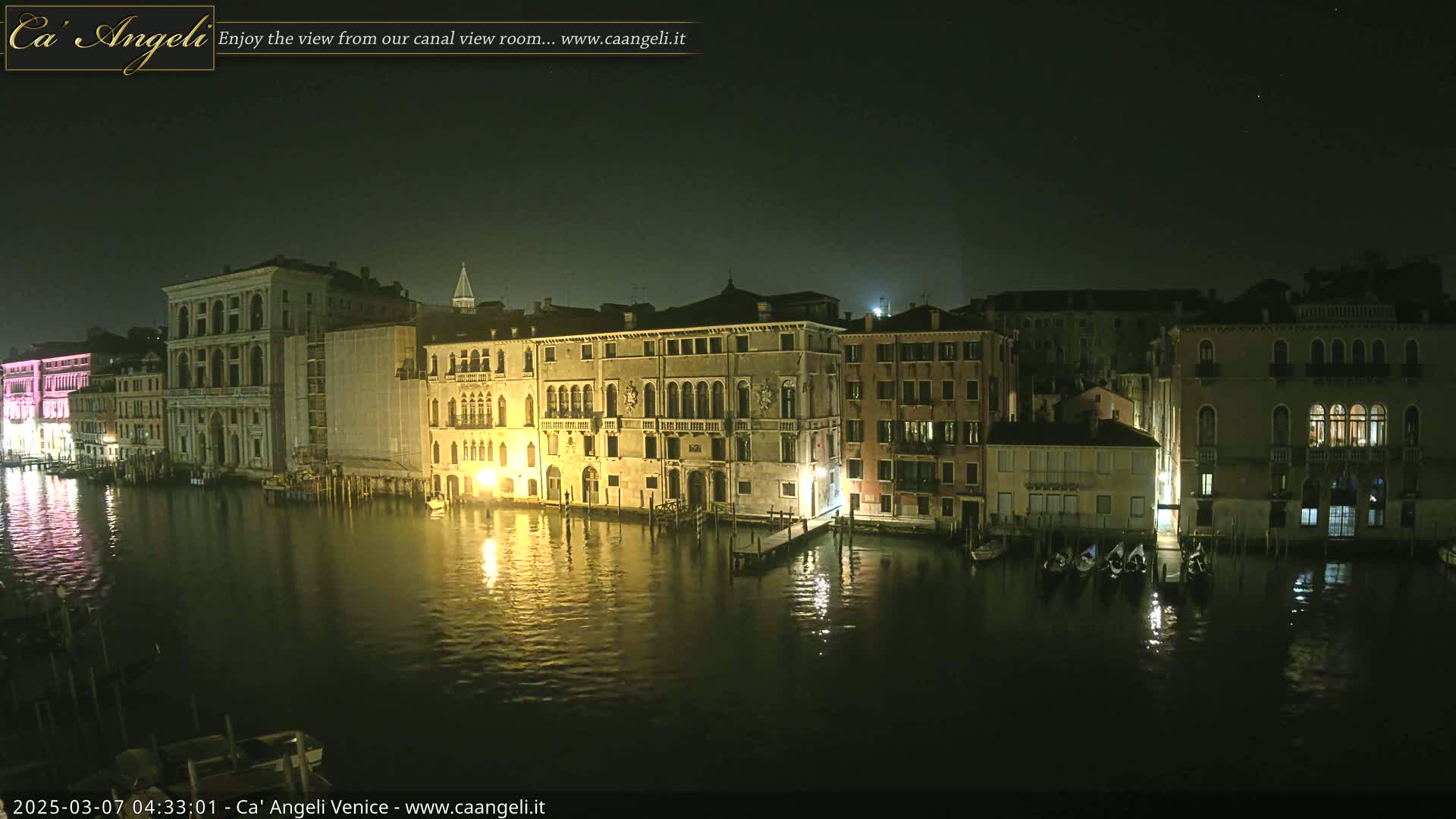 A nighttime view of a canal in Venice, with several illuminated buildings lining the waterway and gondolas moored along the edge.