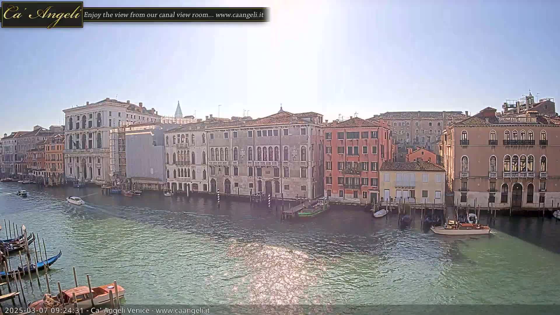 A sunny day in Venice shows a canal lined with historic buildings, gondolas, and boats.
