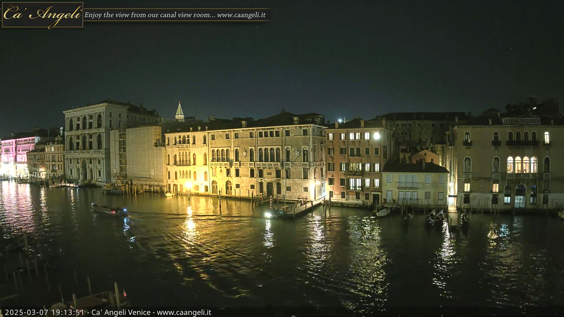 A nighttime view of a canal in Venice, Italy, shows several illuminated buildings lining the waterway, with boats moored along the shore and a few moving on the water under a clear, dark sky.