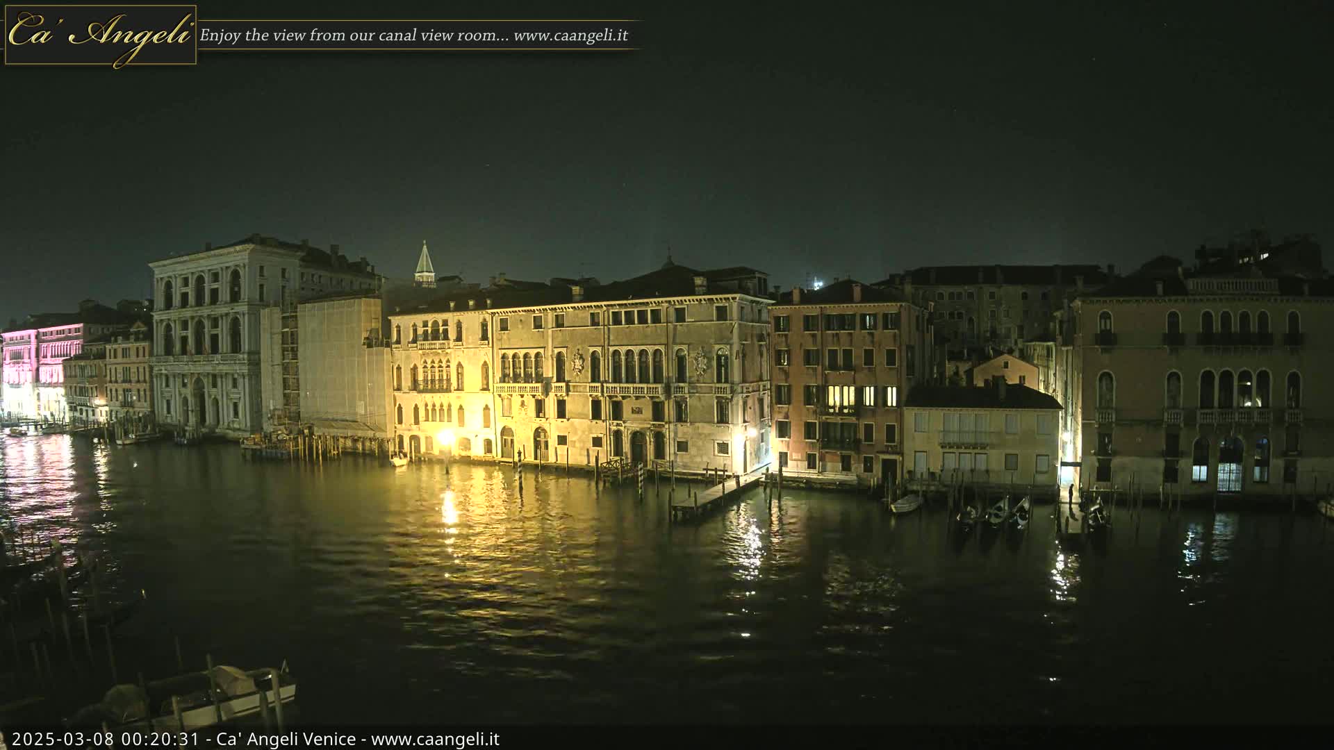A nighttime view of a canal in Venice, Italy, shows several illuminated buildings lining the waterway, with calm water reflecting the lights.