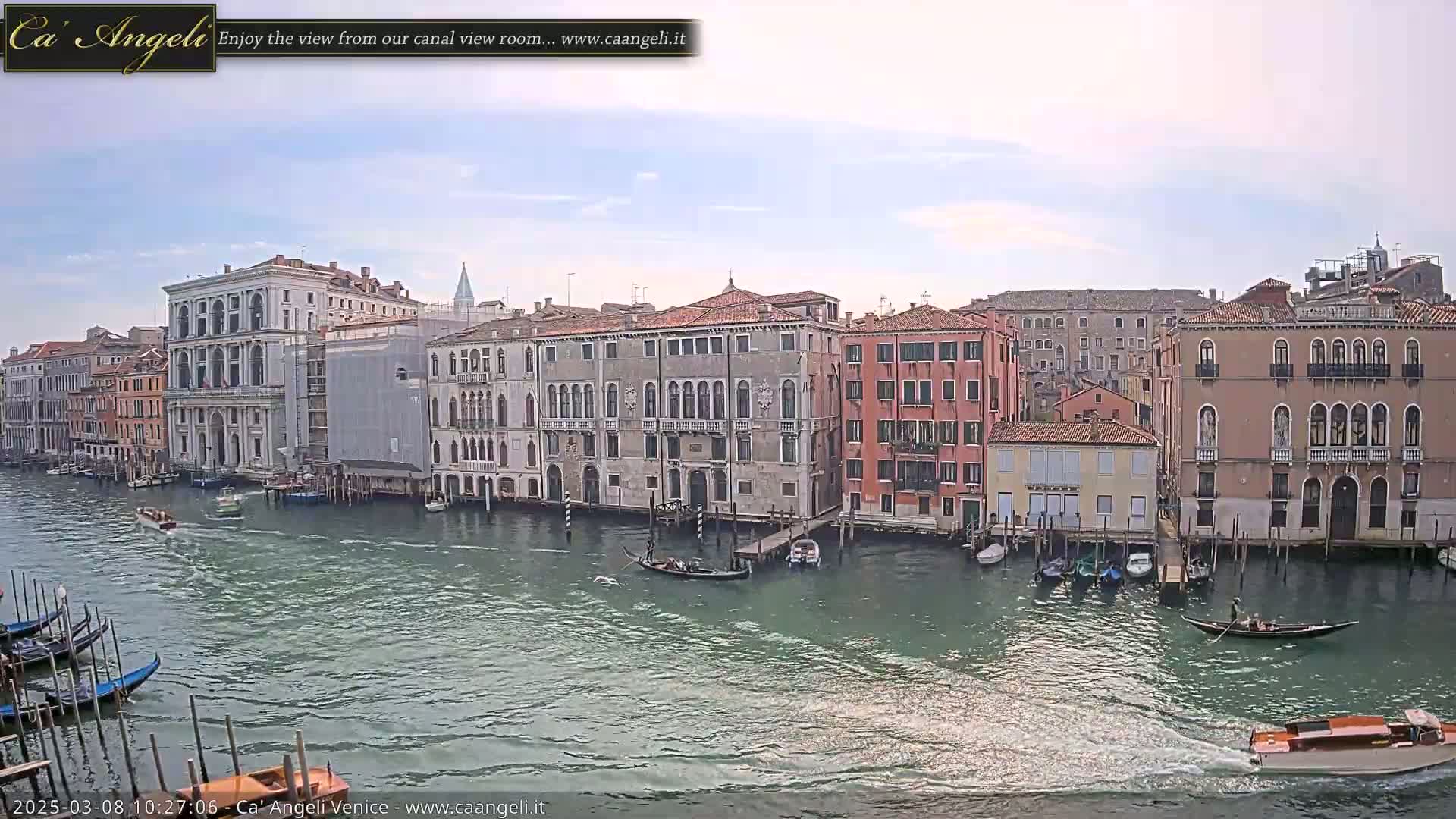 A wide shot shows a waterway with gondolas and other small boats passing by several multi-story buildings under a partly cloudy sky.