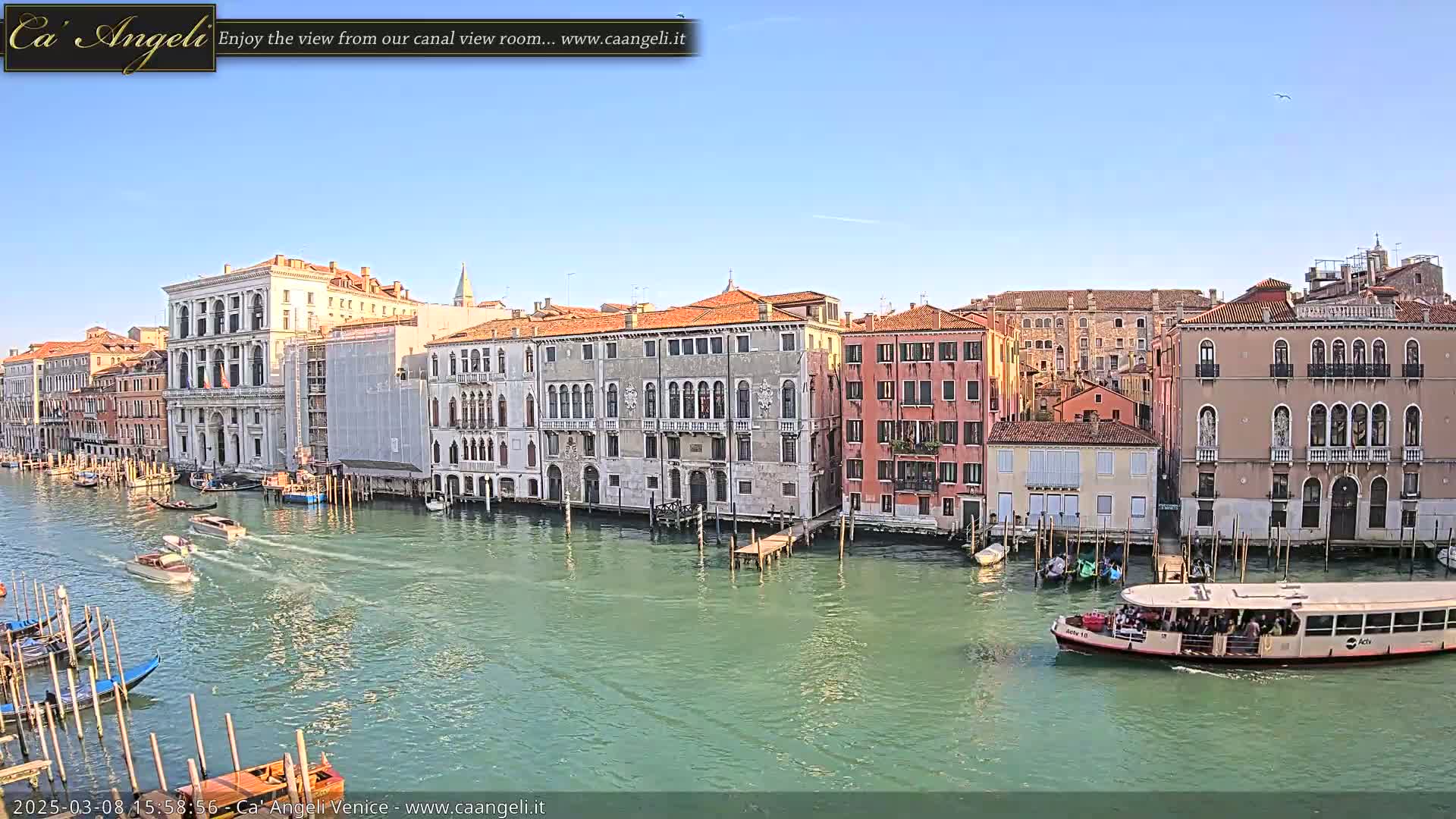 A sunny day in Venice shows a canal with several boats and gondolas passing by historic buildings lining the waterway.