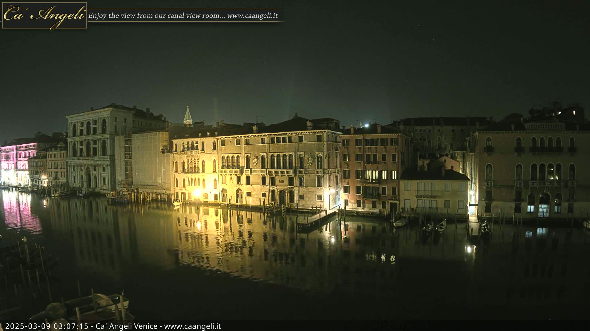 A nighttime view of a calm canal in Venice, with several illuminated buildings lining its banks reflecting in the dark water under a clear, starry sky.