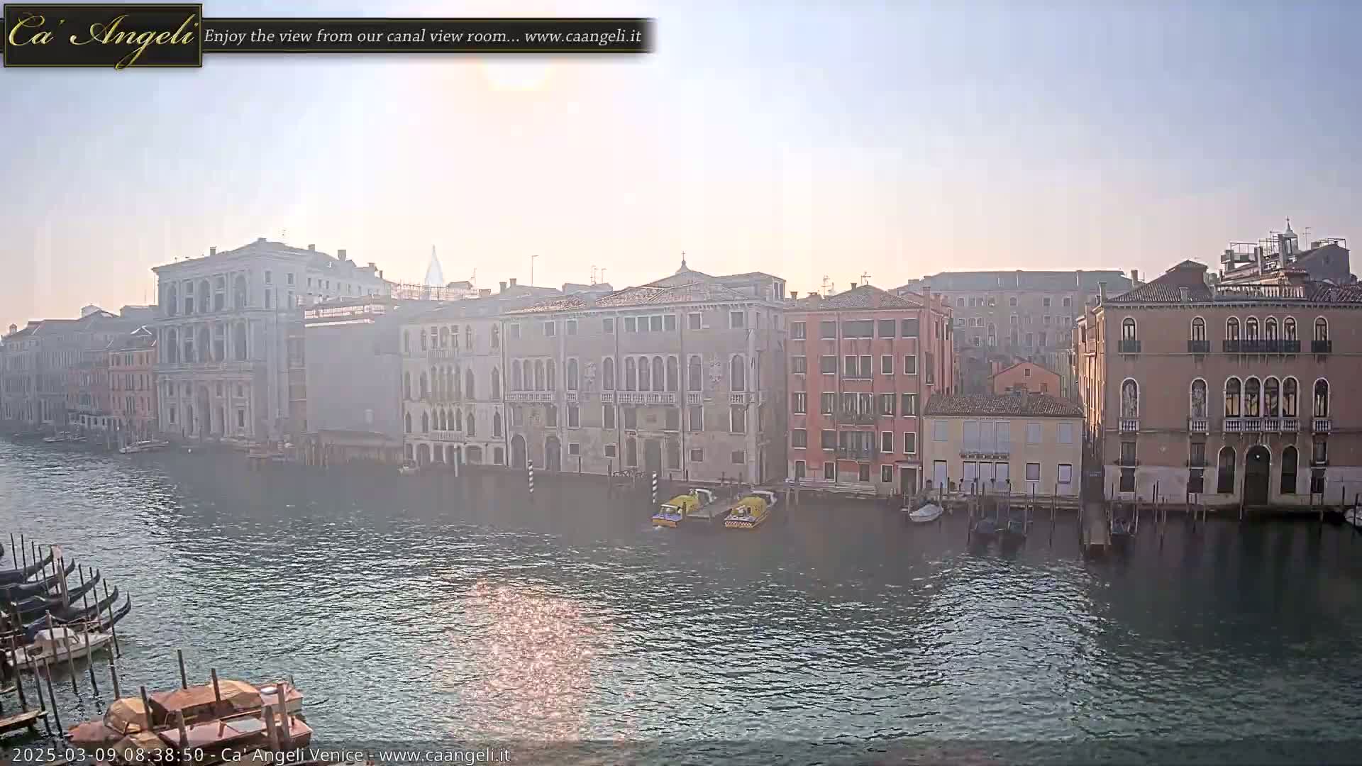 A hazy sunrise illuminates a Venetian canal, where several buildings line the waterway, with gondolas and water taxis moored along the banks.