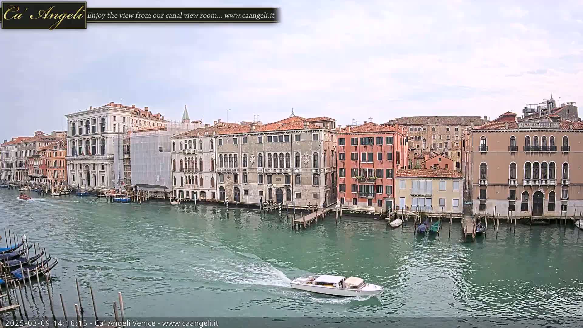 A motorboat speeds across a calm canal in Venice, past several gondolas moored along the banks lined with historic buildings under an overcast sky.
