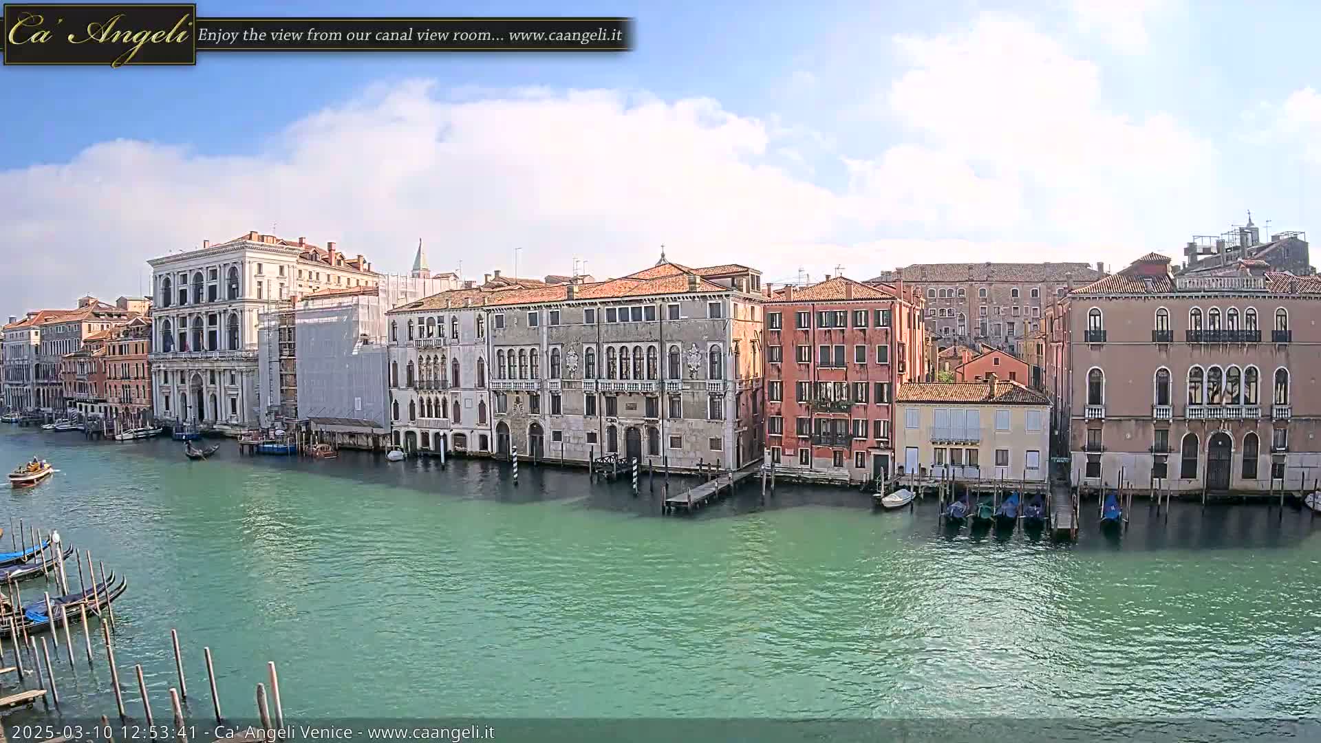 A sunny day in Venice shows several buildings lining a canal with gondolas moored along the waterfront.