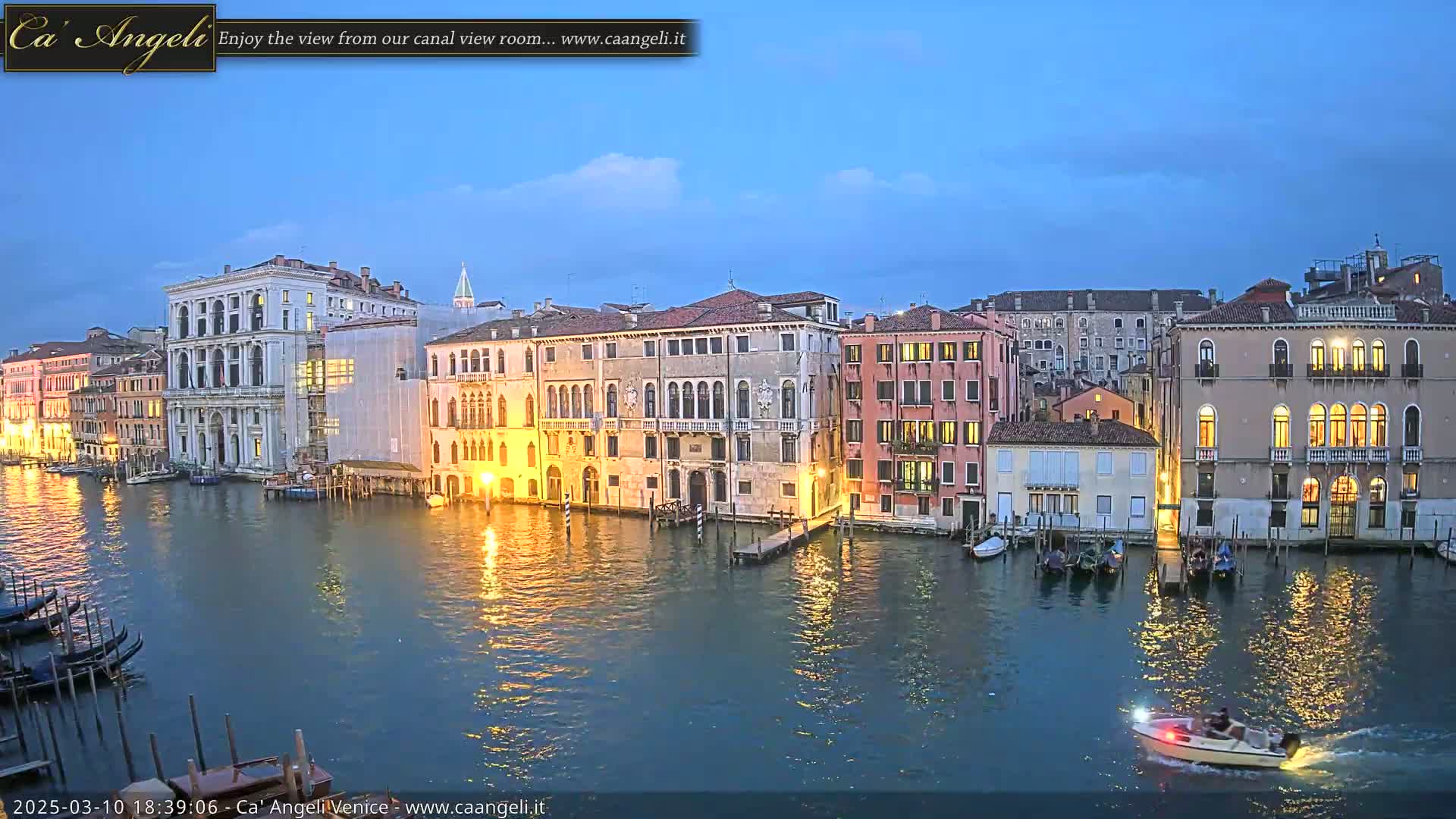 A twilight view of a canal in Venice, with numerous buildings illuminated along the waterway, and a small motorboat traveling on the calm water.