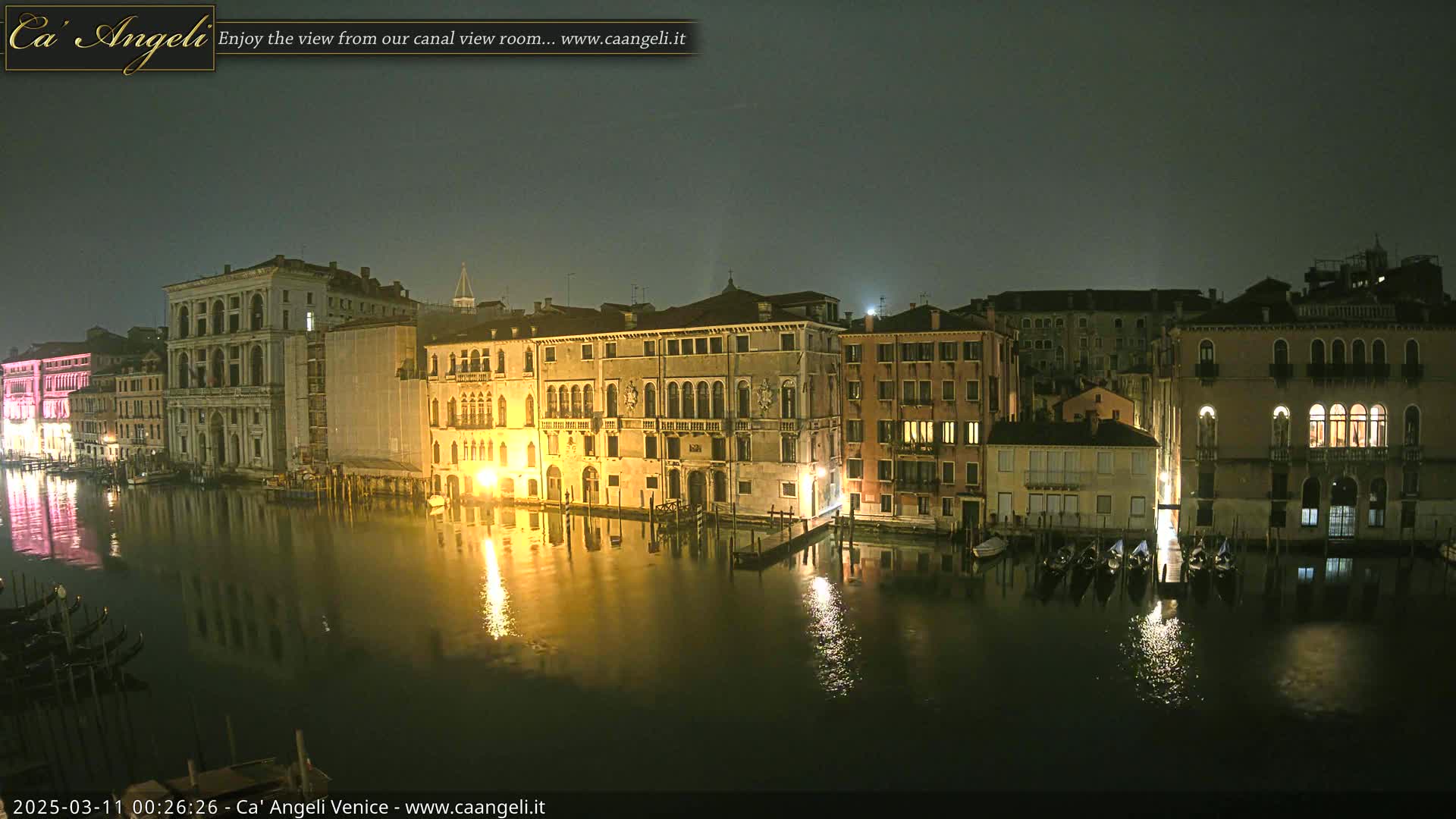 Several illuminated buildings line a calm canal at night under a dark, possibly overcast sky.