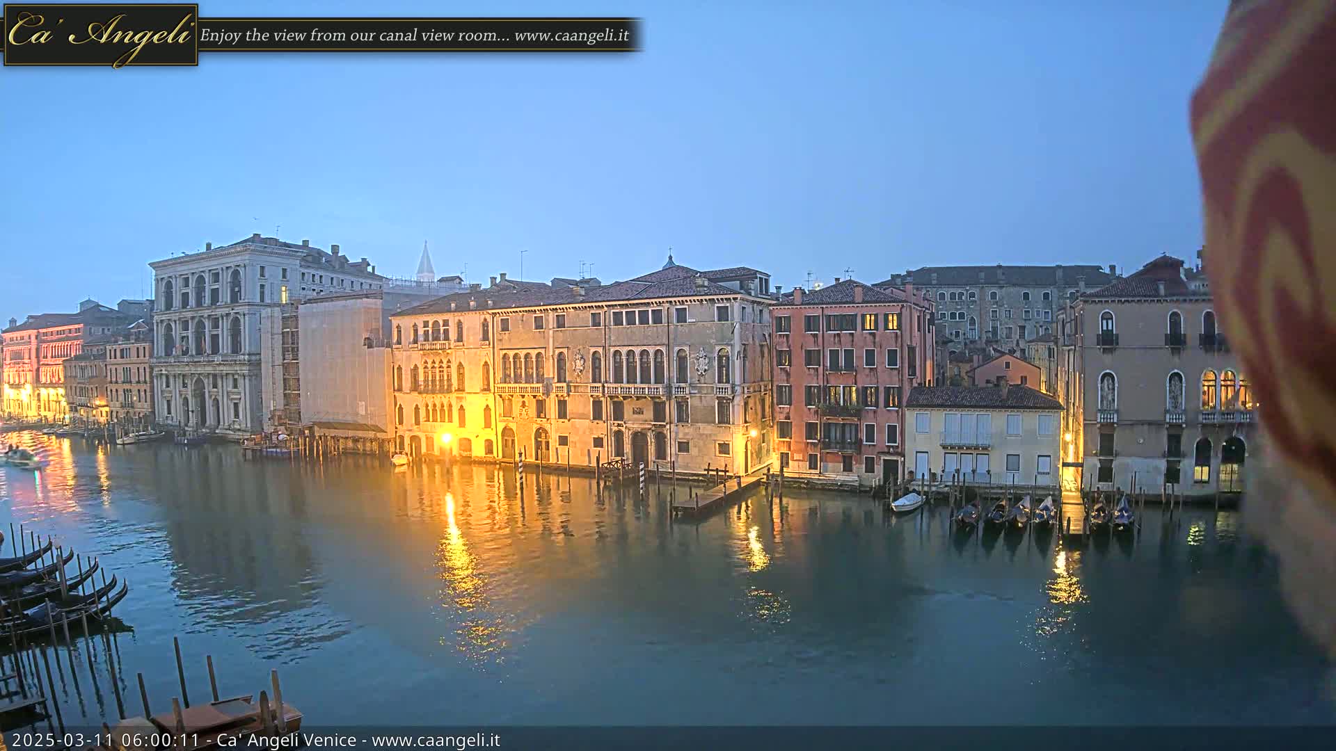 A twilight view shows a canal in Venice, Italy, with several gondolas moored along the waterway and buildings lining the banks, their lights reflecting on the calm water.
