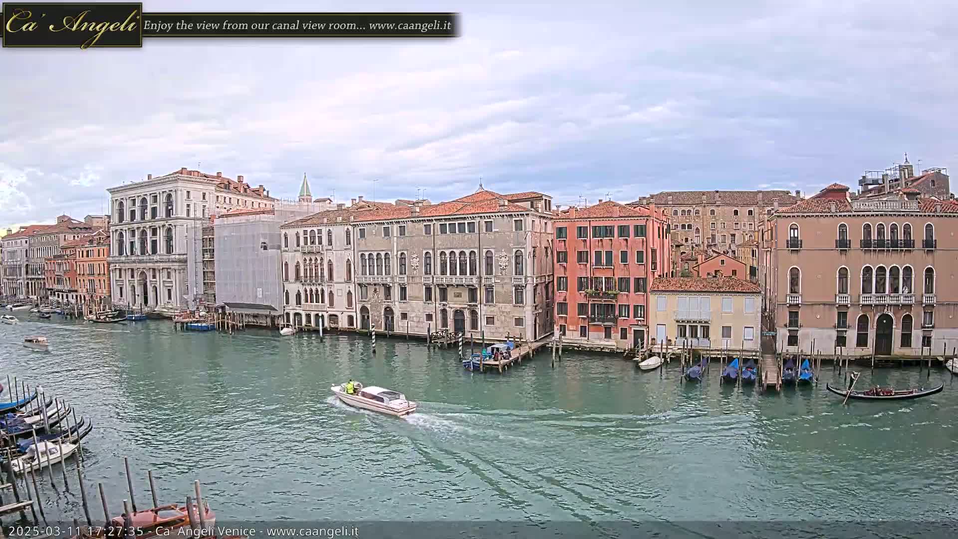 A motorboat travels down a canal in Venice, Italy, under a mostly cloudy sky, passing by numerous buildings and moored gondolas.