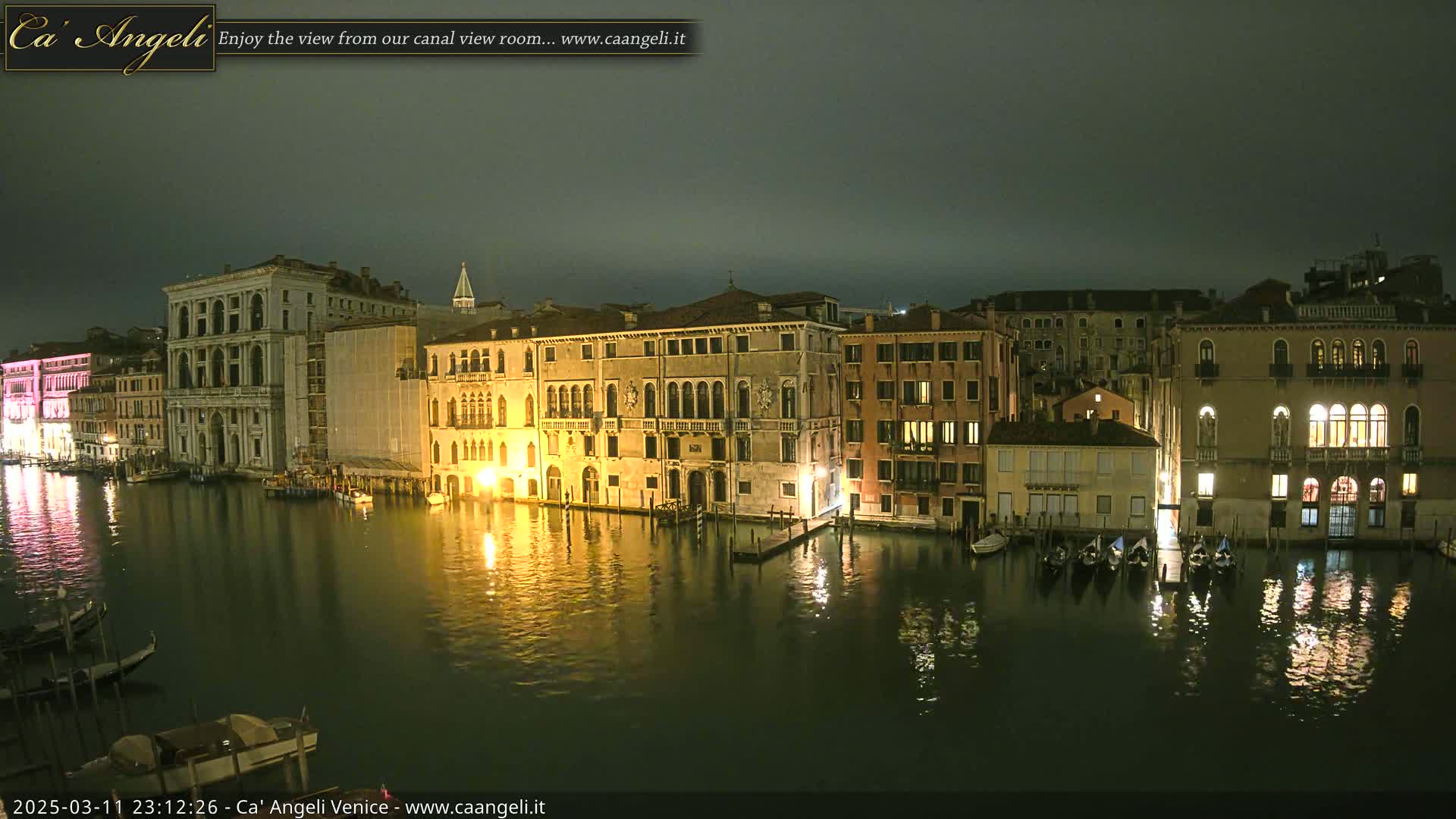A nighttime view of a canal in Venice, Italy, showing several illuminated buildings along the waterfront and gondolas moored alongside.