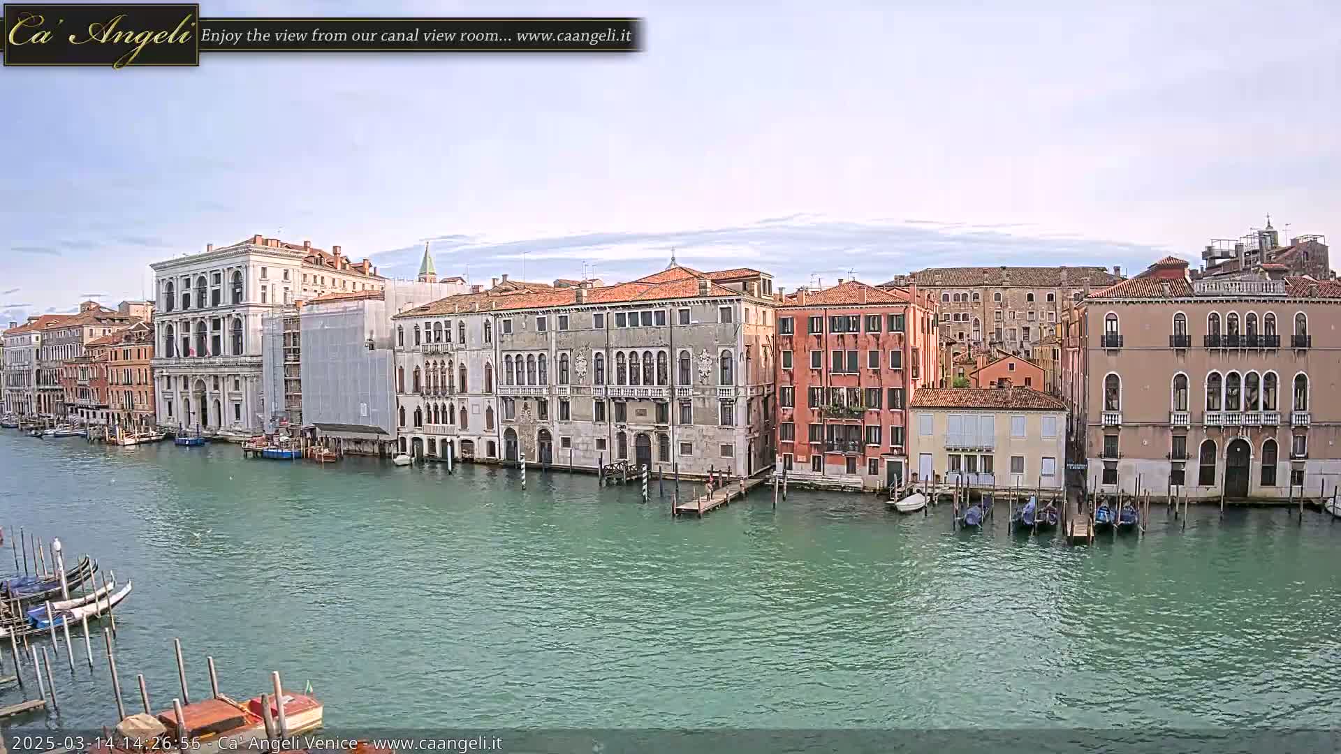 A wide shot of a calm canal in Venice, Italy, shows several historic buildings lining the waterway, with gondolas moored along the shore under a mostly clear sky.