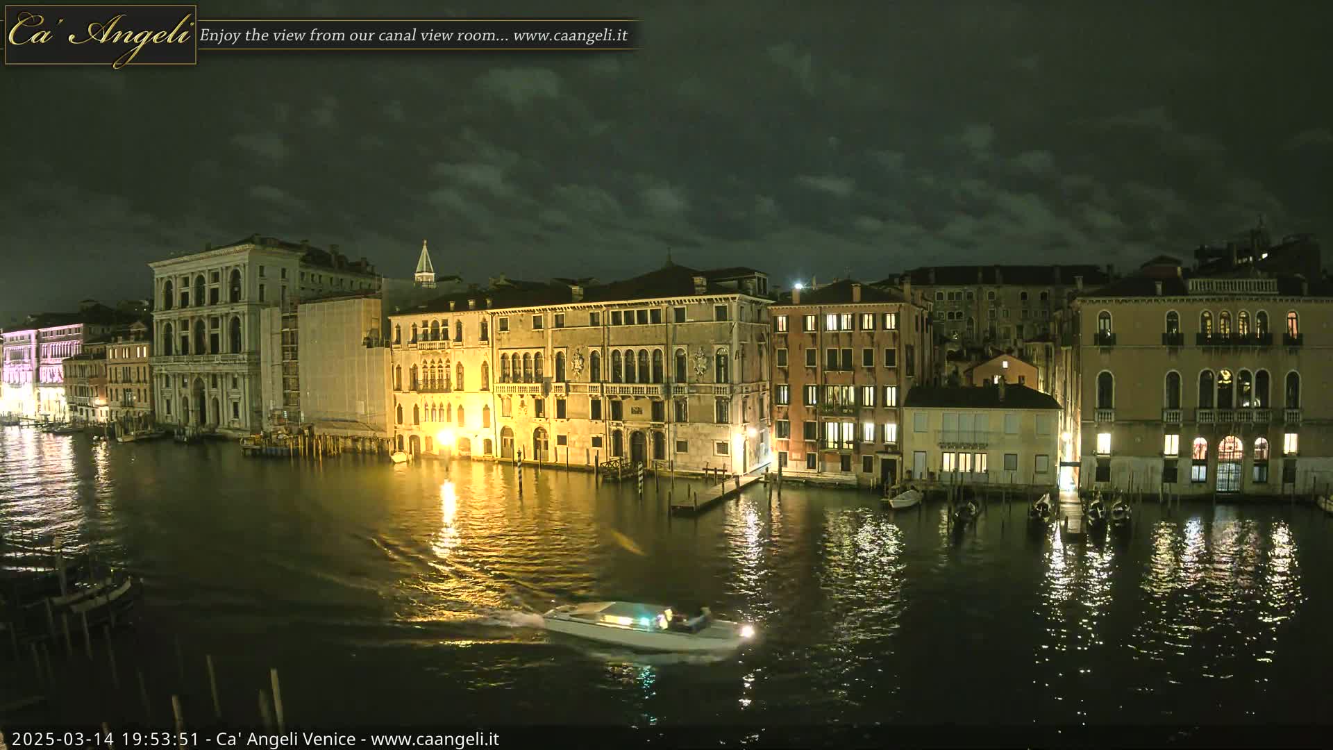 A nighttime view of a canal in Venice, under a partly cloudy sky, shows several illuminated buildings along the water's edge with a small motorboat moving across the water.