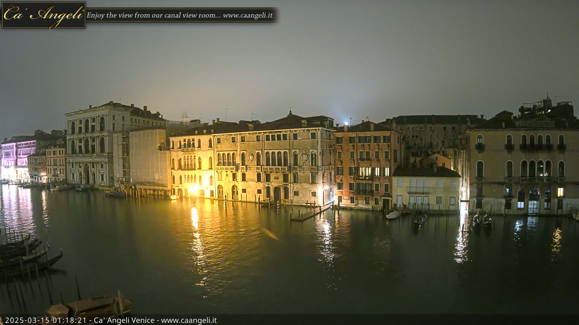 A nighttime view of a canal in Venice, showing several illuminated buildings along the waterway under a foggy sky.