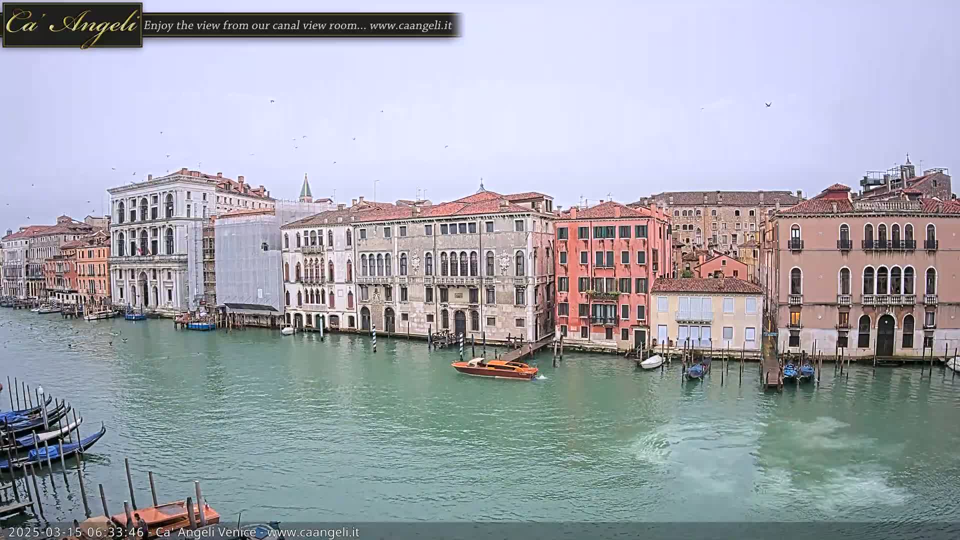 A canal in Venice, Italy, on an overcast day, shows several historic buildings lining its banks, with gondolas and a motorboat on the water.