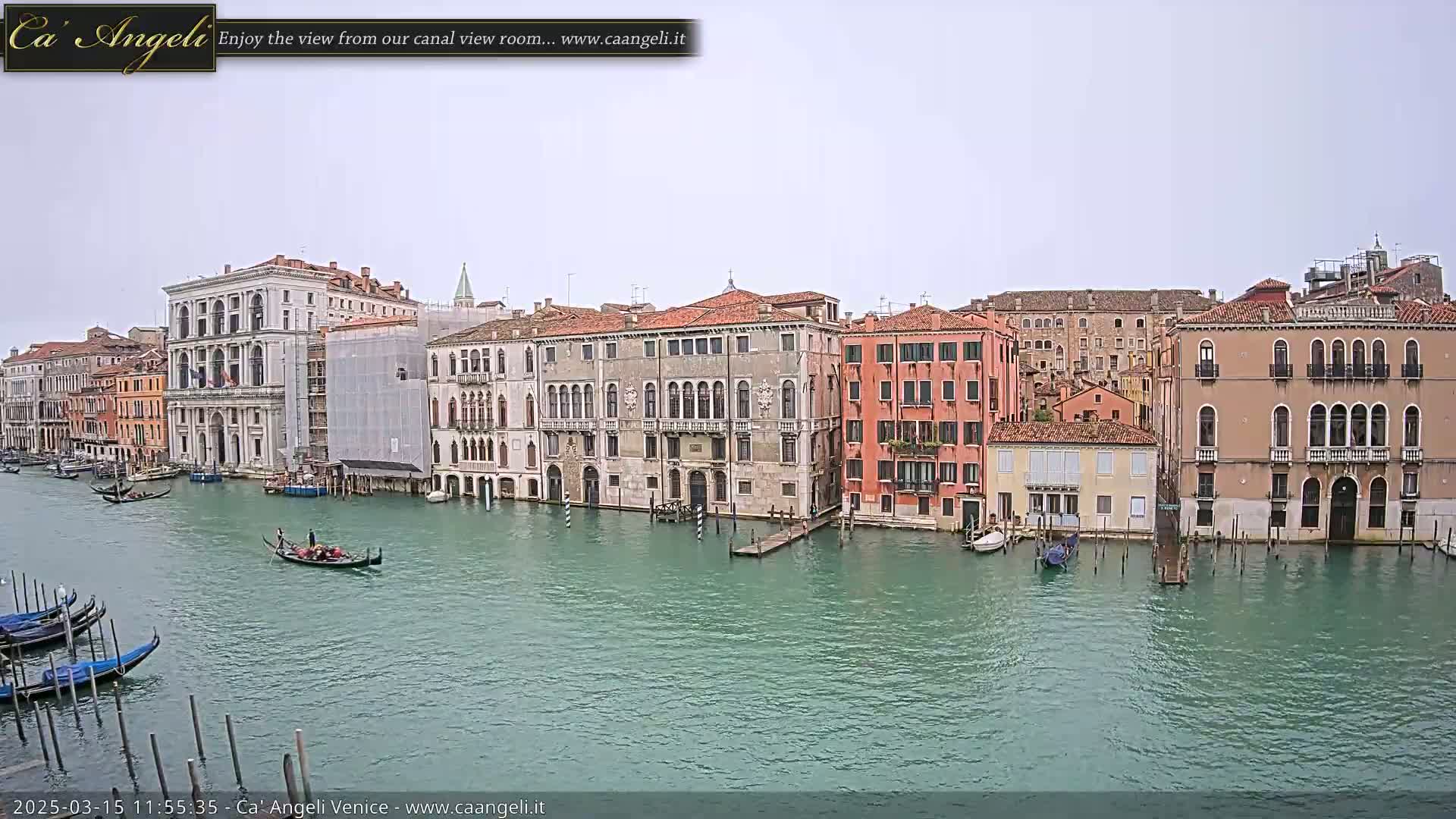 A wide shot of a Venetian canal on an overcast day shows several gondolas and numerous buildings lining the waterway, one of which is under construction.