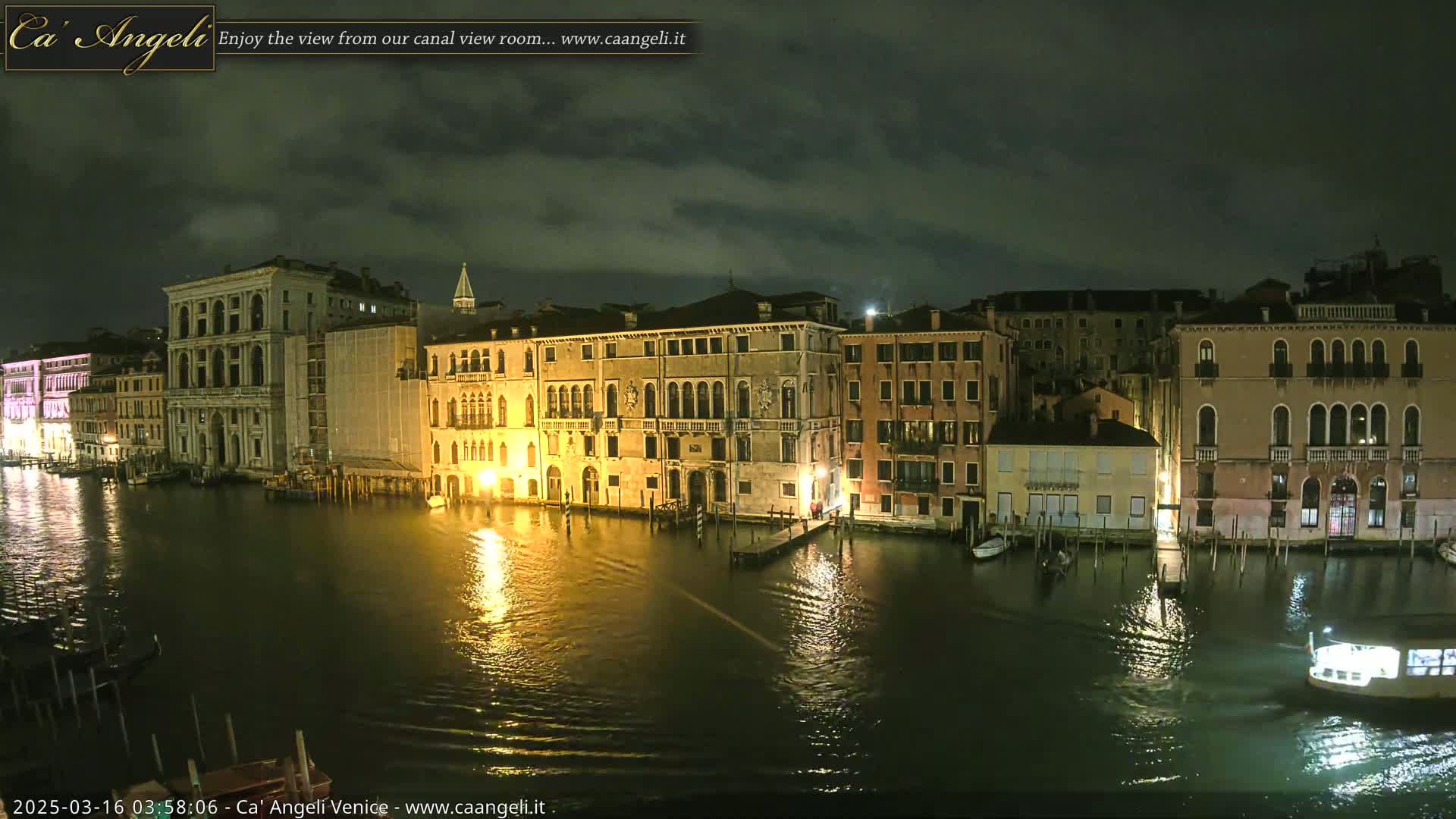 A nighttime view of a canal in Venice, under a cloudy sky, shows illuminated buildings lining the waterway, with a boat visible on the water.