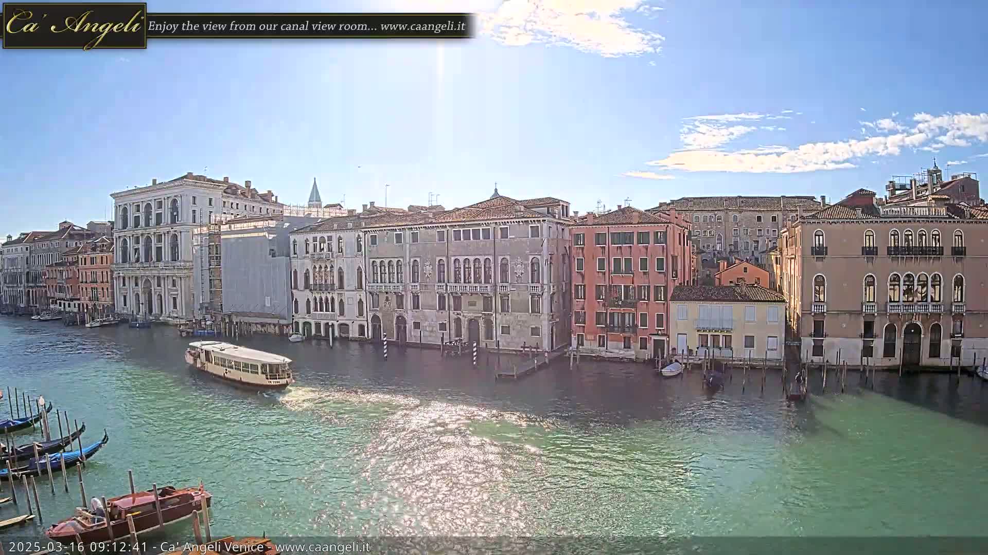 A sunny day in Venice shows a canal with gondolas, a water bus, and numerous buildings lining the waterway.