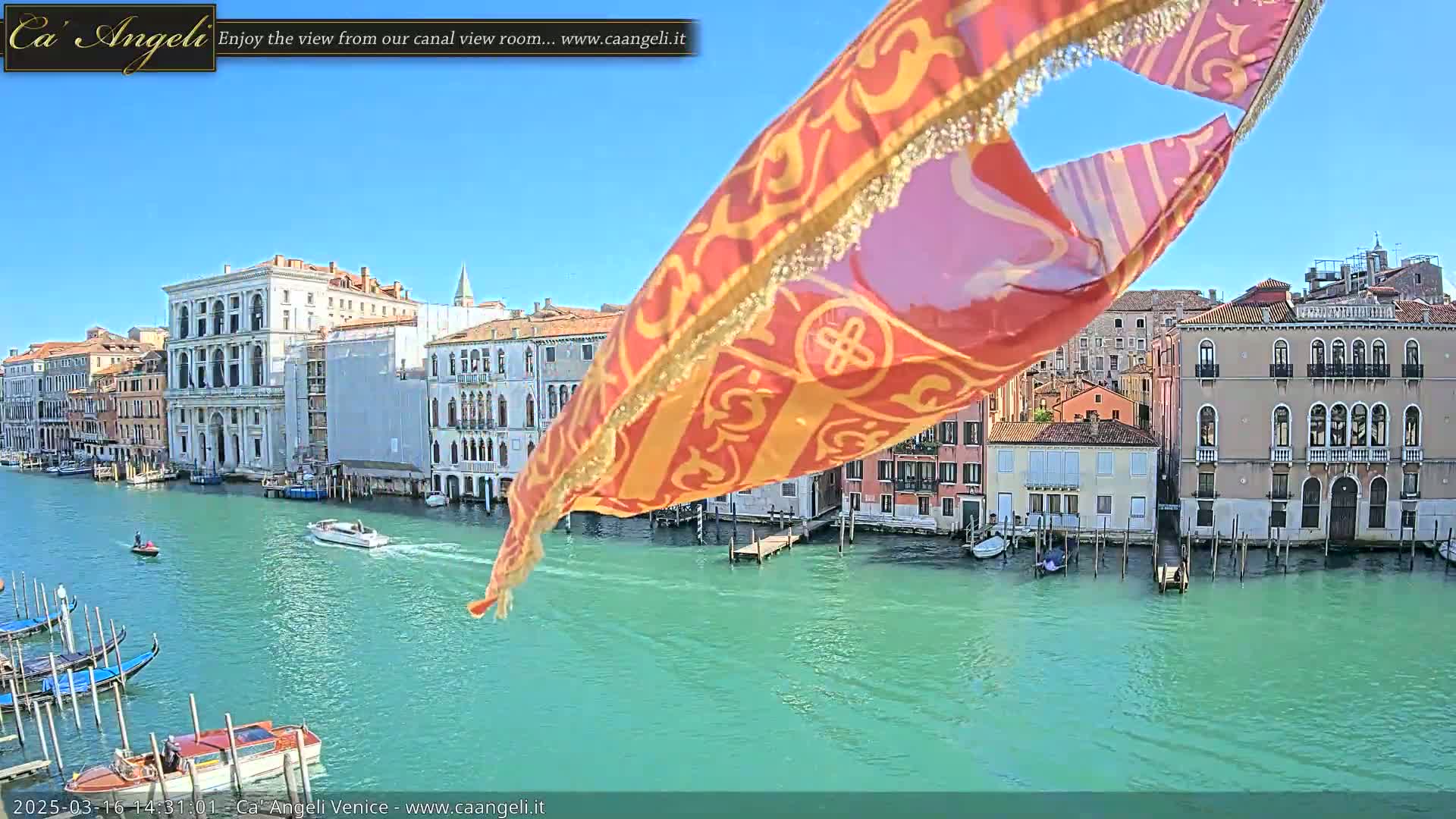A colorful flag partially obscures a view of a Venetian canal on a sunny day, with boats moored along the waterway and buildings lining the banks.