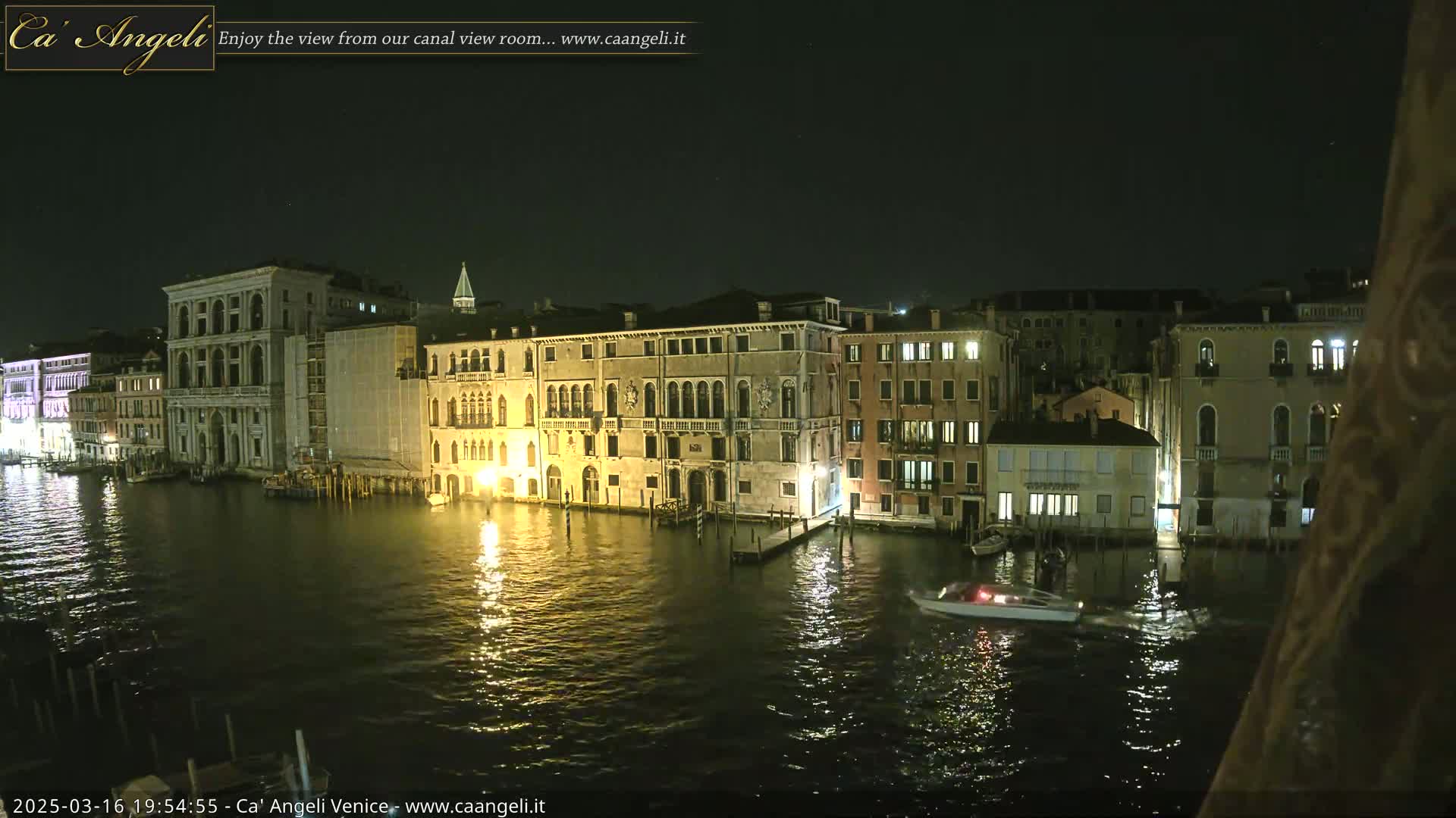 A nighttime view of a canal in Venice, Italy, with several illuminated buildings lining the waterway, and a small boat traveling on the calm water.