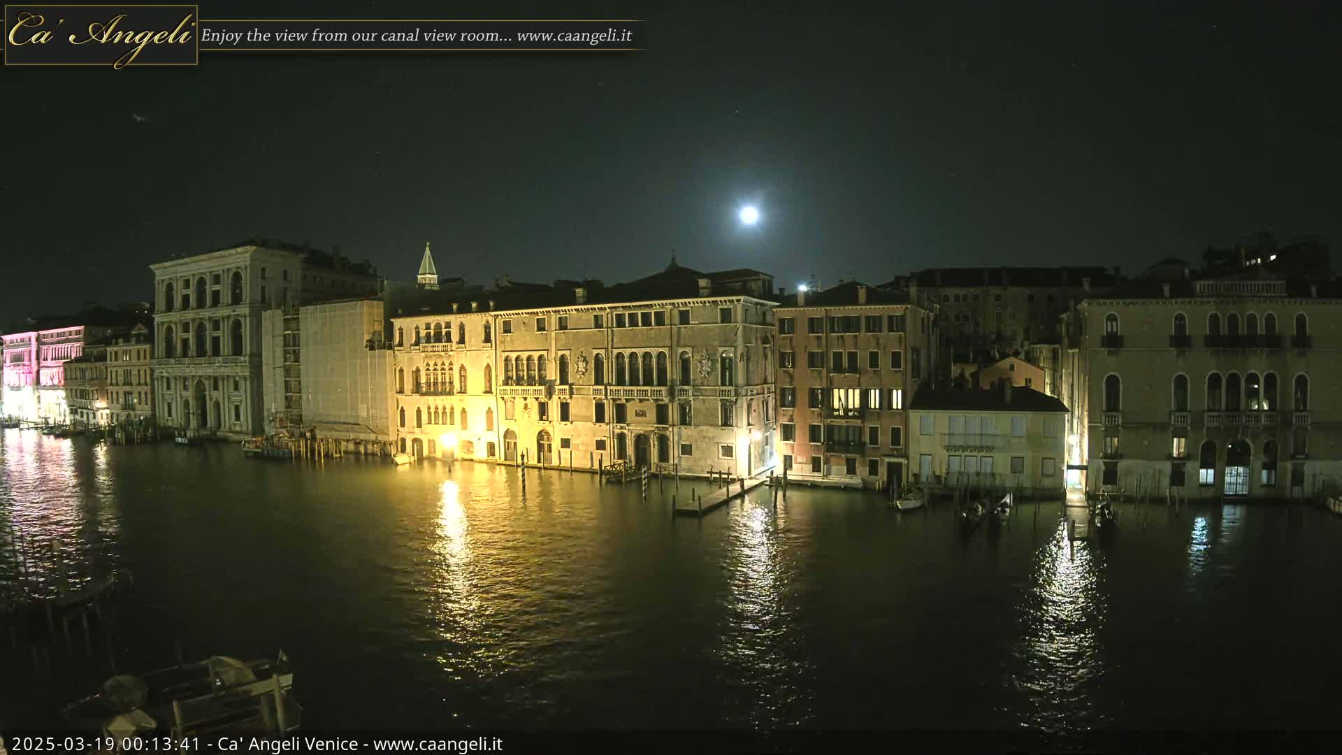 A moonlit night scene shows a canal in Venice, with several buildings illuminated along its banks, their lights reflecting in the dark water.