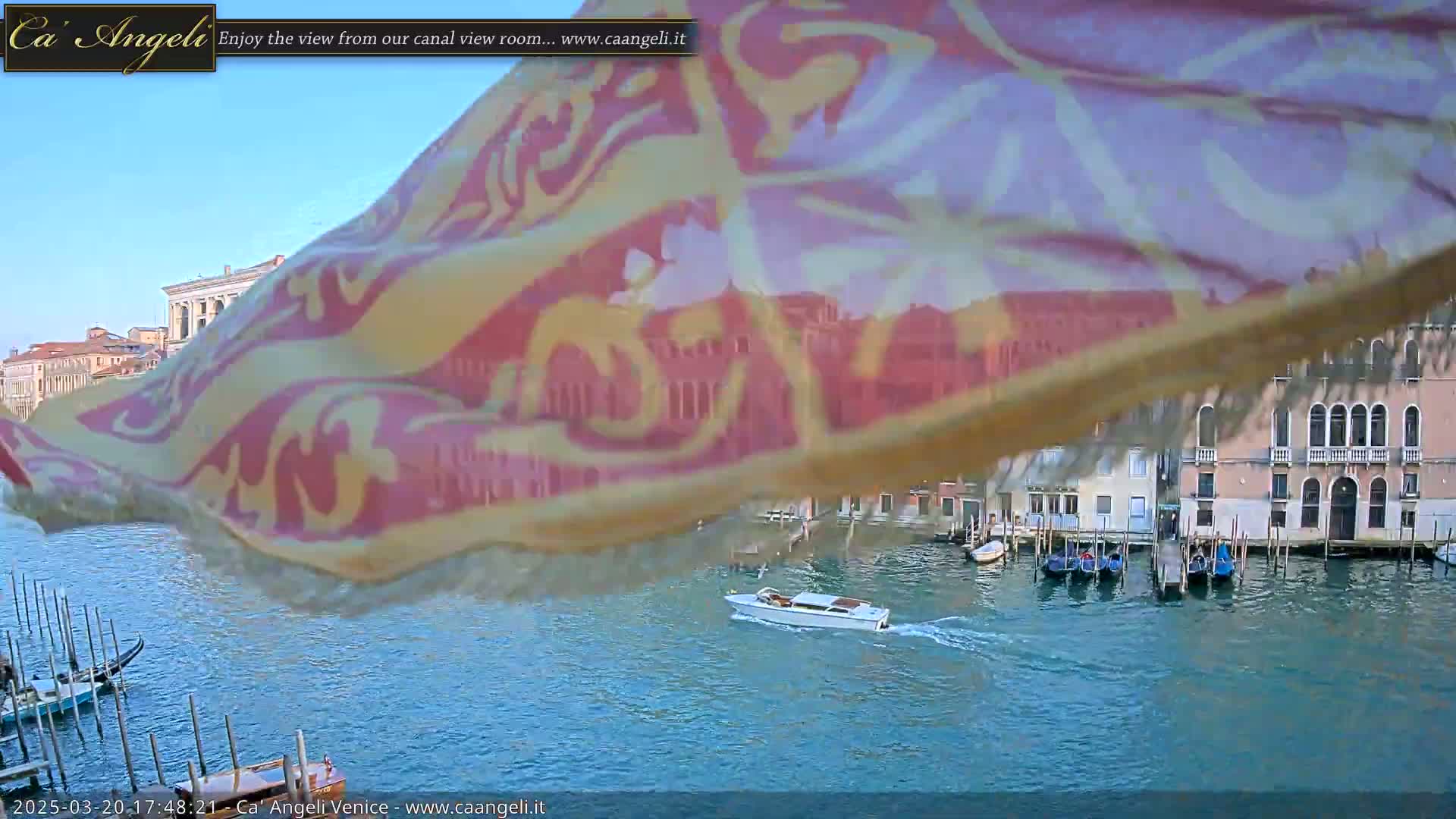A patterned fabric billows in the wind over a Venetian canal scene on a clear day, with several gondolas and a motorboat visible on the water.