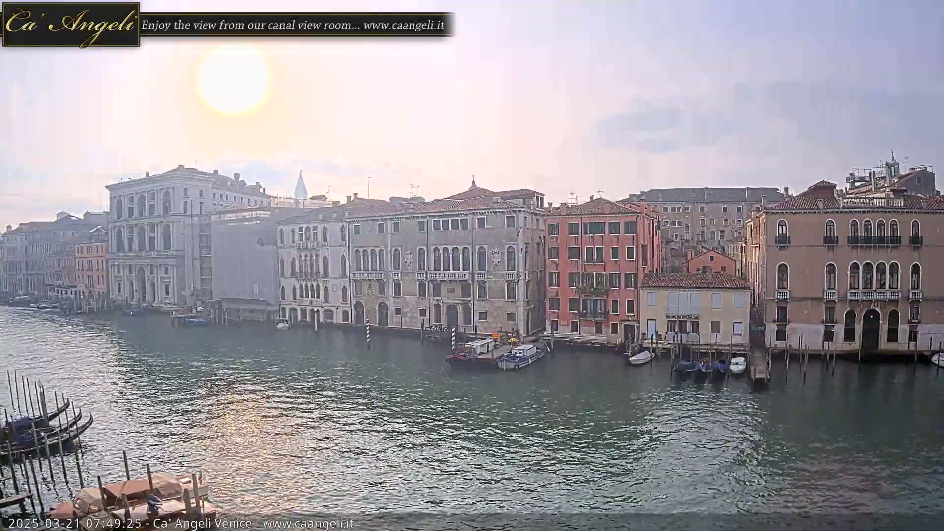 A hazy sunrise illuminates a Venetian canal lined with historic buildings and moored boats.