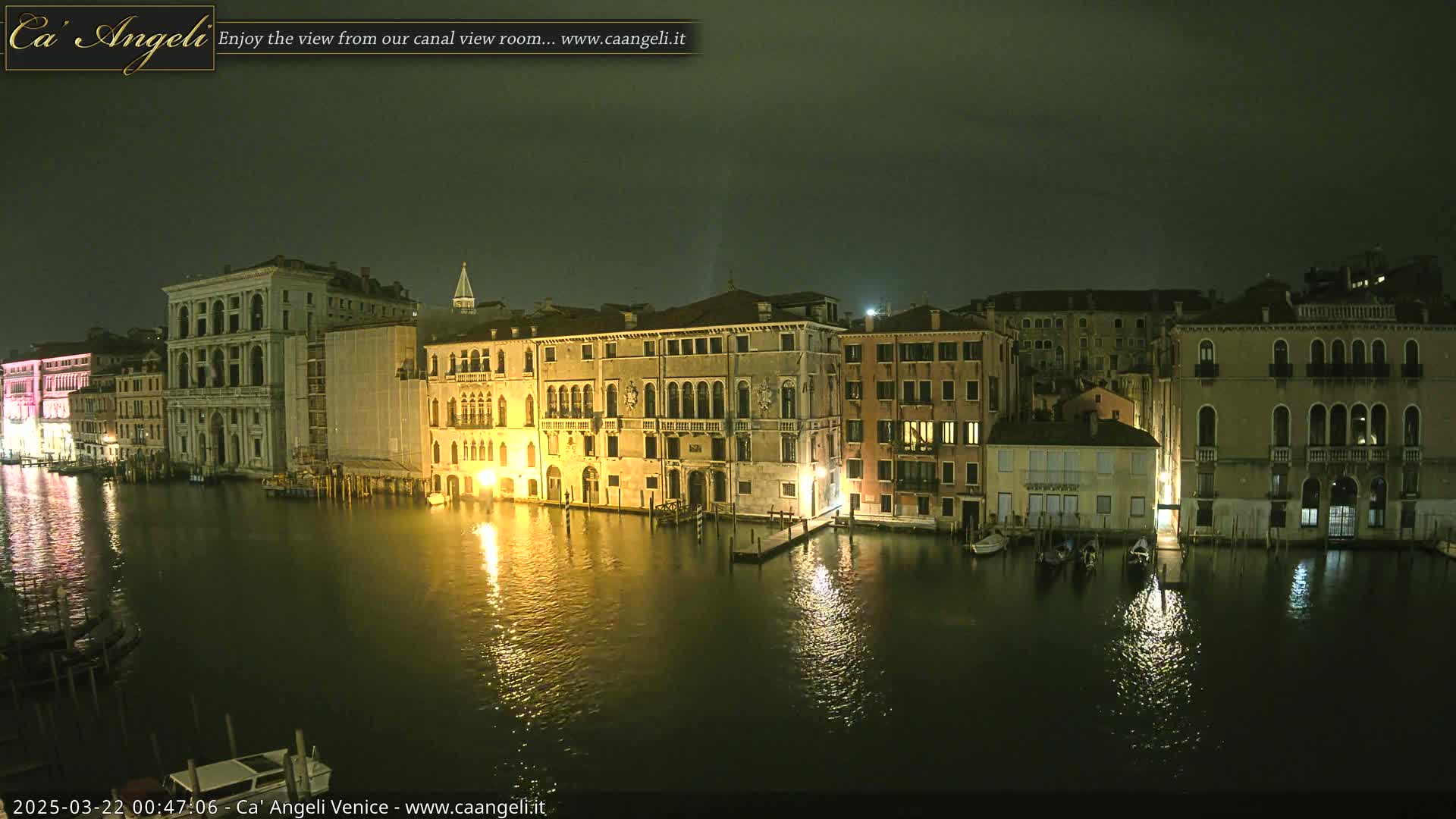 A nighttime view of illuminated buildings along a calm canal in Venice under a dark, overcast sky.