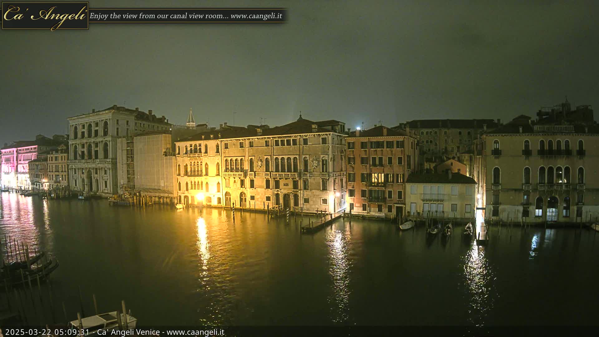 A nighttime view of a canal in Venice, with several buildings lit along the water's edge under a cloudy sky.