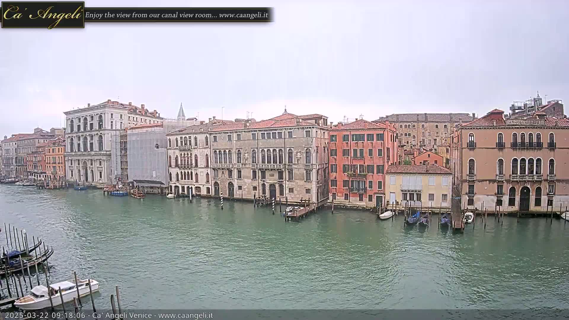 A wide shot of a canal in Venice on an overcast day, showing several multi-story buildings along the waterway with gondolas and other small boats moored alongside.