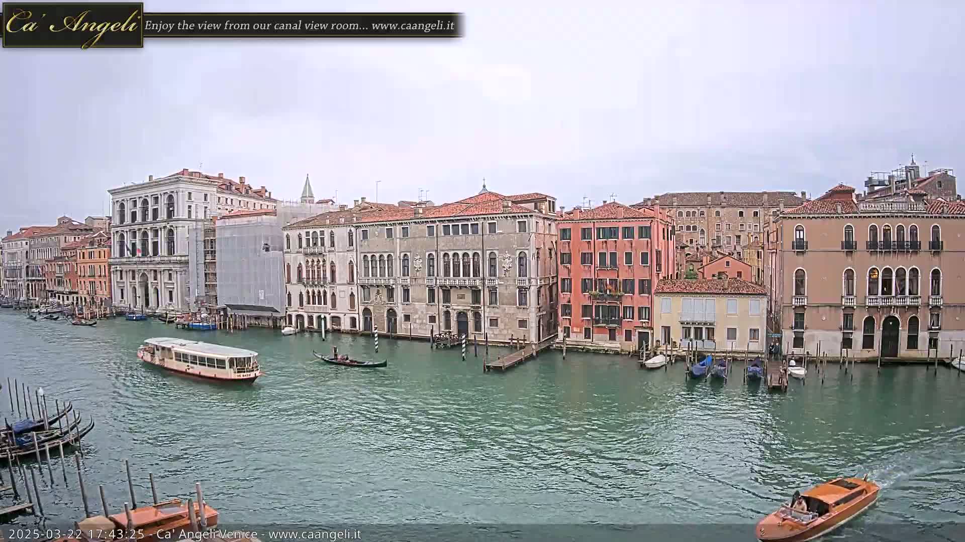 A waterway in Venice, Italy, shows several gondolas and water taxis passing by tall, colorful buildings on an overcast day.