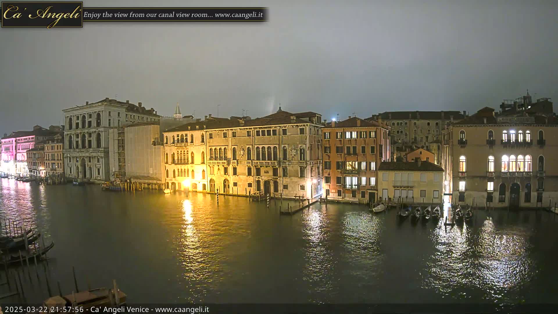 A nighttime view of a canal in Venice, Italy, under a cloudy sky, shows several illuminated buildings lining the waterway, with gondolas moored along the shore.