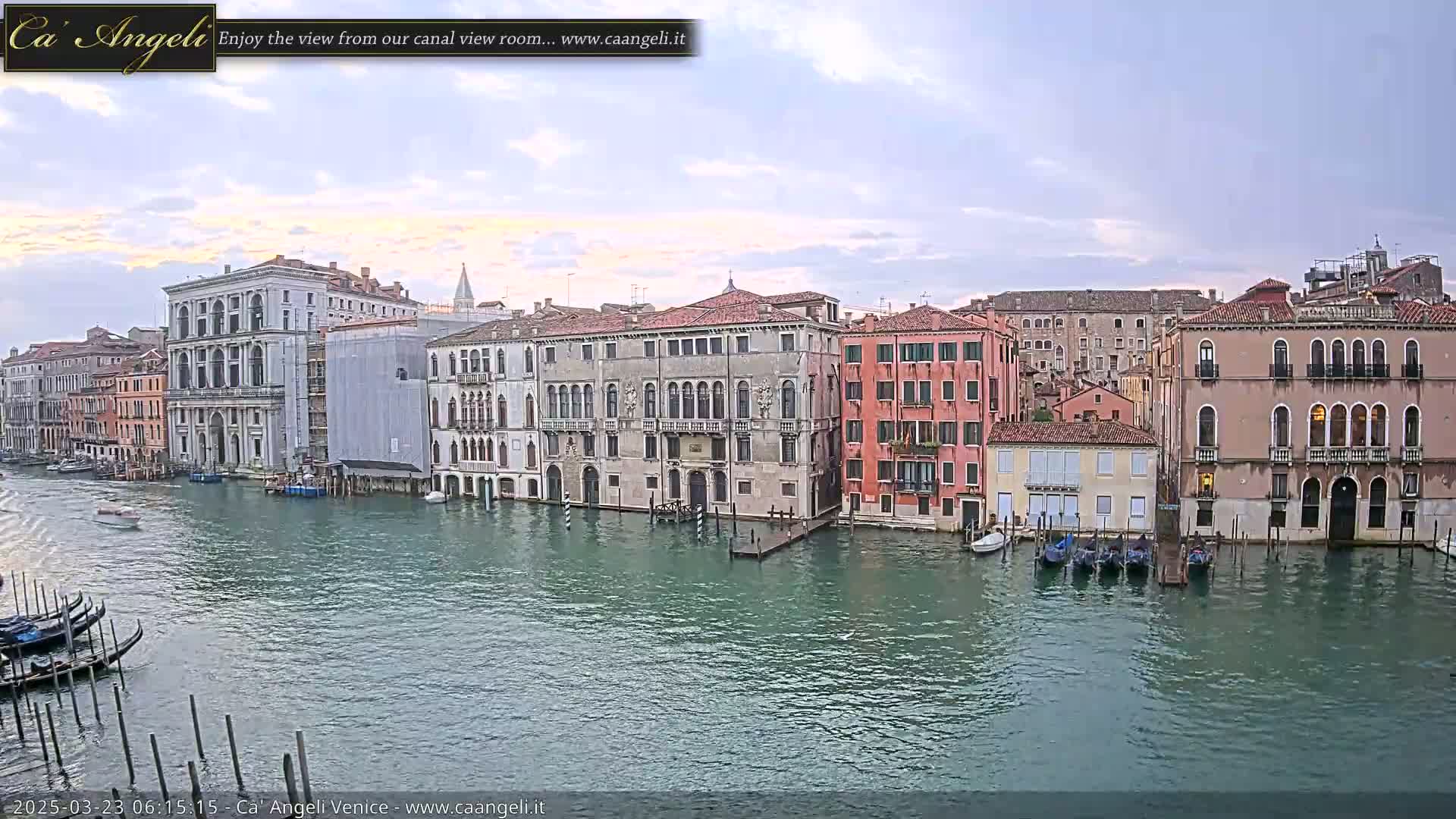 A calm canal in Venice, under a partly cloudy sky, is lined with several historic buildings and moored gondolas.