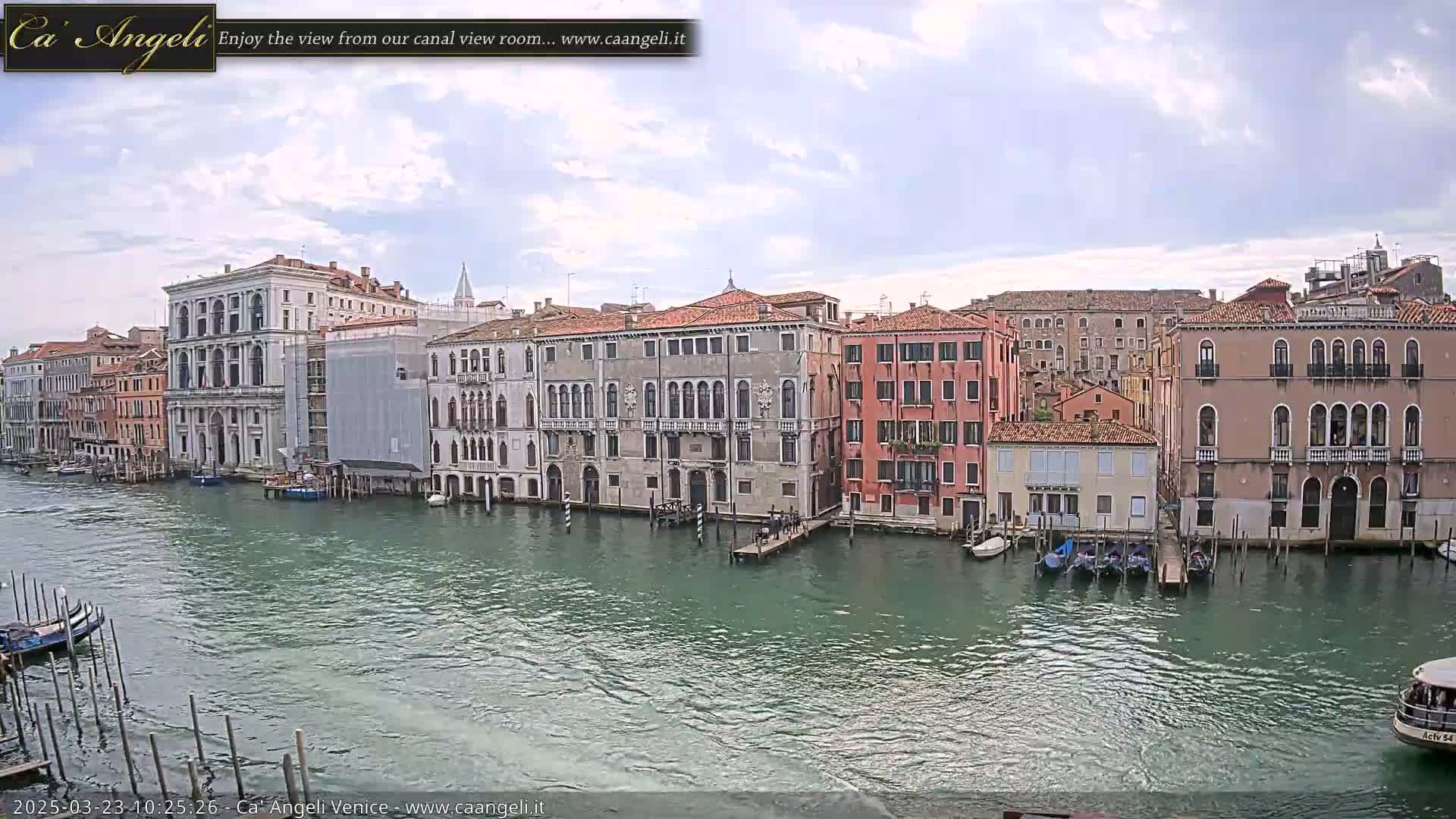 A canal in Venice, under a partly cloudy sky, is lined with historic buildings and several moored boats.