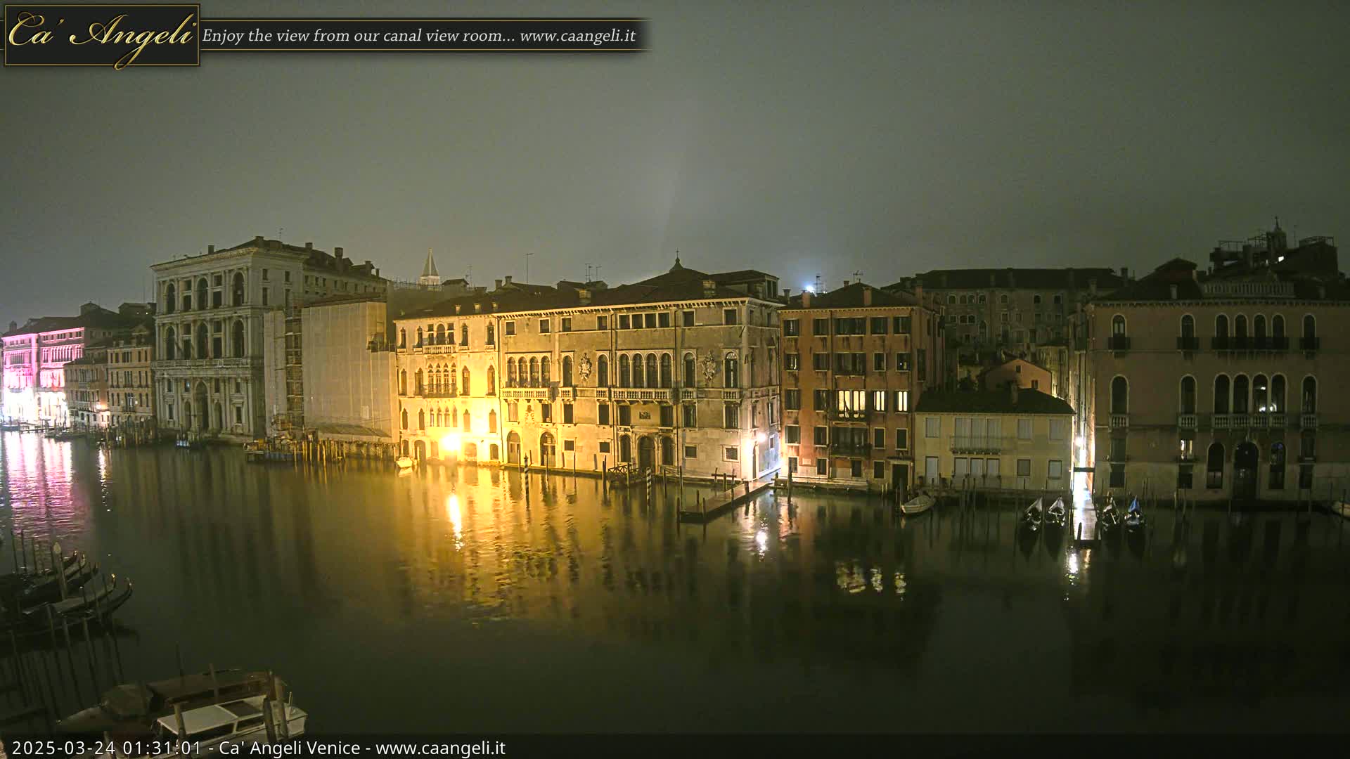 A nighttime view of illuminated buildings lining a canal in Venice under a hazy sky.