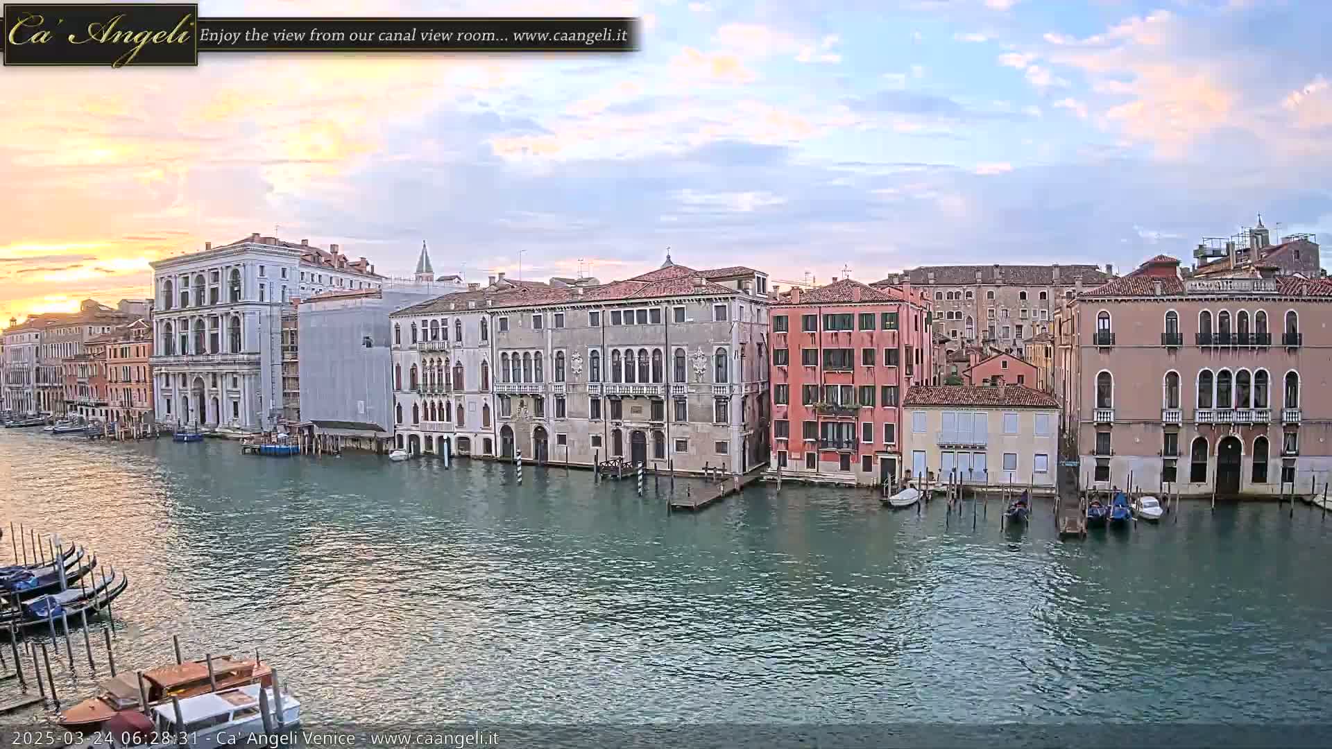 A calm canal in Venice at sunrise, lined with historic buildings and a few moored boats.