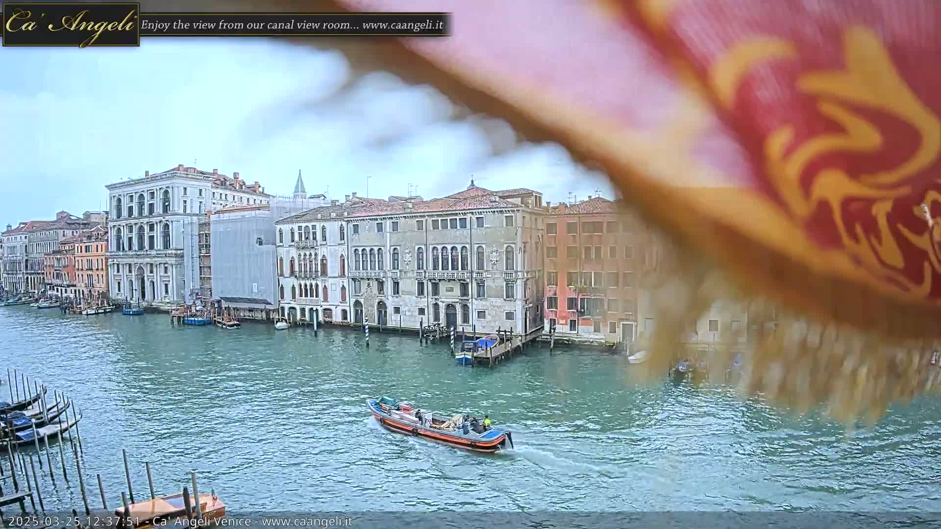 A canal in Venice, under an overcast sky, is shown with several buildings lining its sides, a small boat in the water, and a portion of a patterned fabric obscuring the upper right corner.