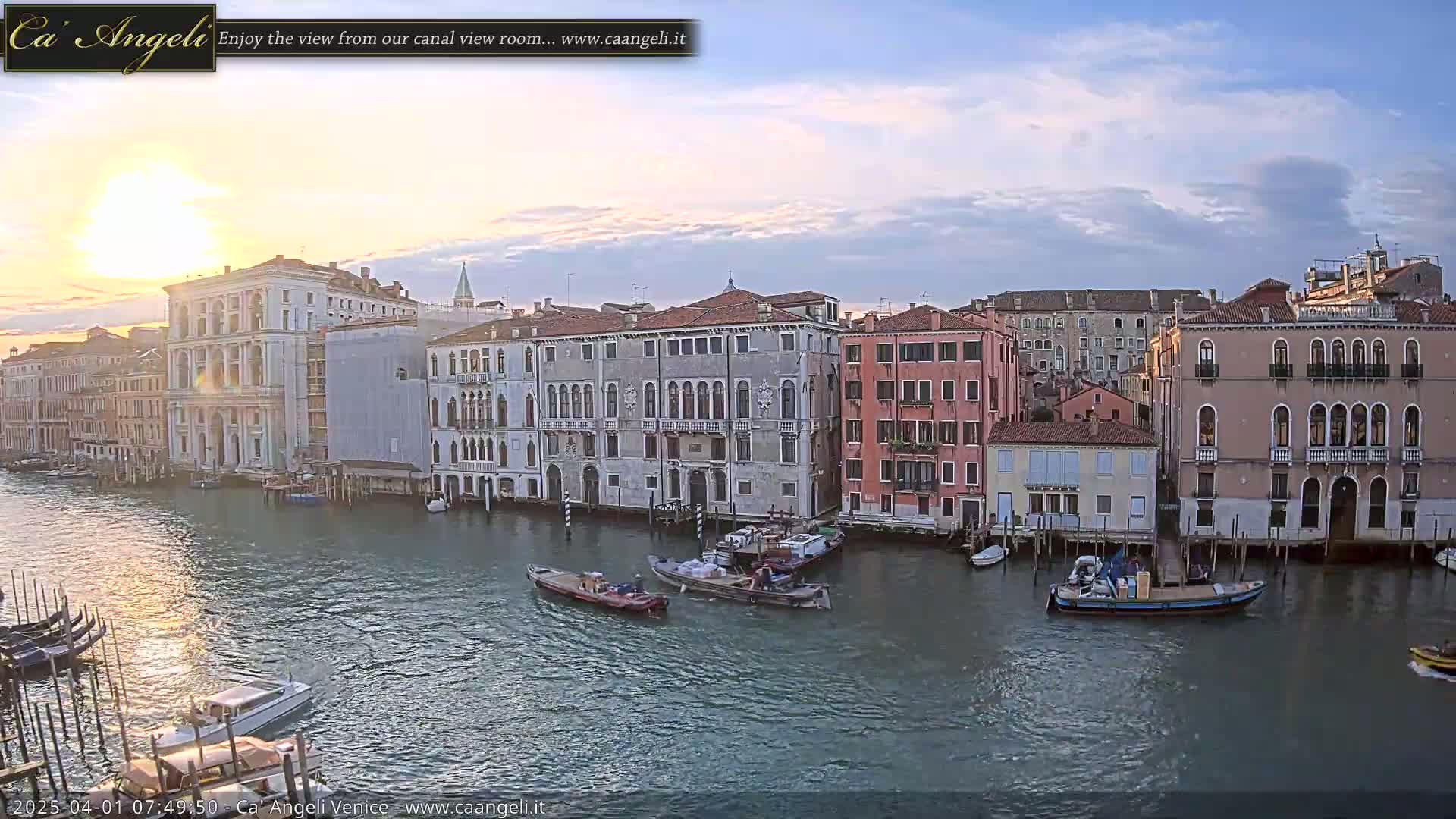 A sunrise illuminates a calm canal in Venice, where several boats are moored alongside historic buildings.