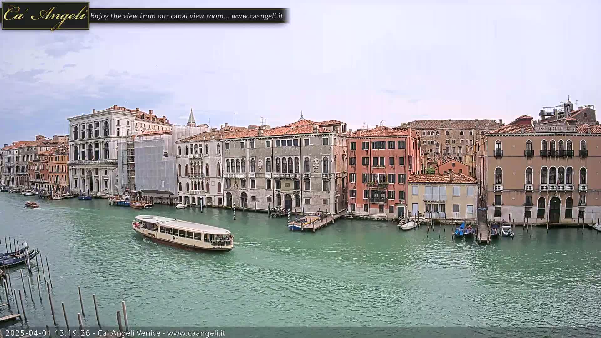 A waterway in Venice, under a mostly cloudy sky, shows a variety of buildings along its banks with boats and gondolas moored nearby, and a water bus traveling on the canal.