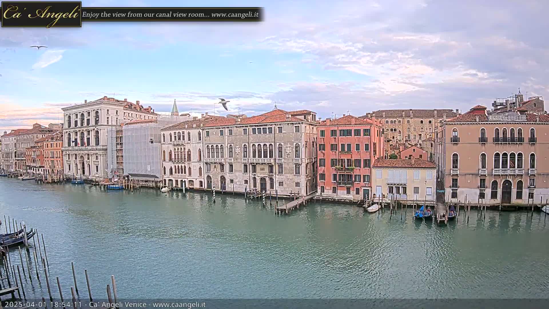 A canal in Venice, Italy, is shown with several buildings lining its banks under a partly cloudy sky.