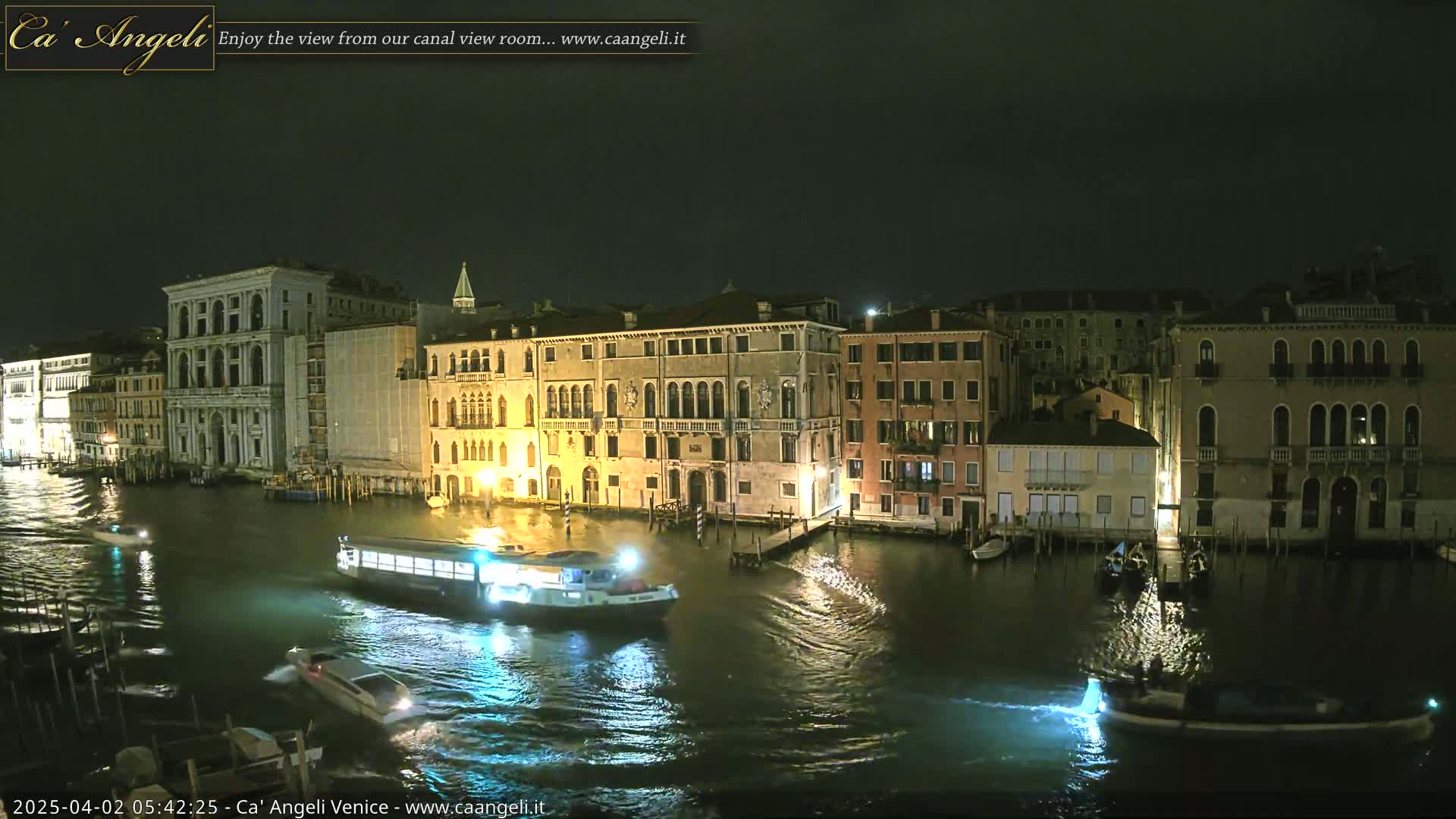 A nighttime view of a canal in Venice, Italy, shows several buildings lit along the waterway with boats moving on the water.