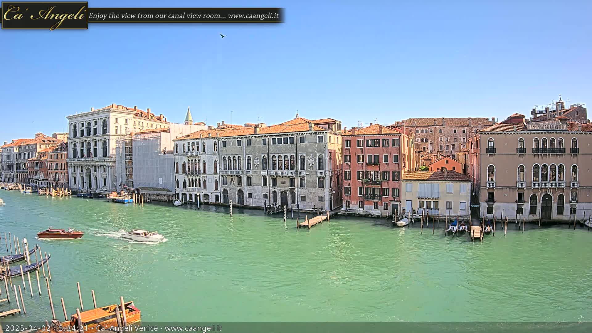 A waterway with green water is lined with several multi-storied buildings under a clear blue sky; several boats, including gondolas, are on the water.