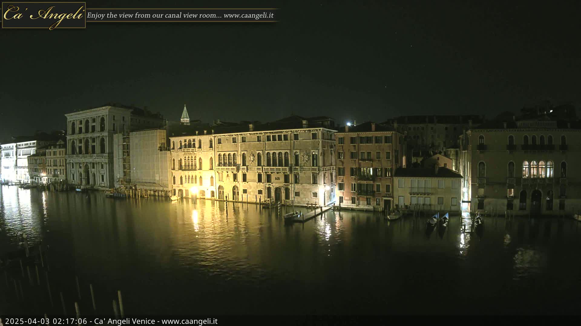 A nighttime view of several illuminated buildings lining a calm canal, with gondolas moored along the waterfront under a clear, dark sky.