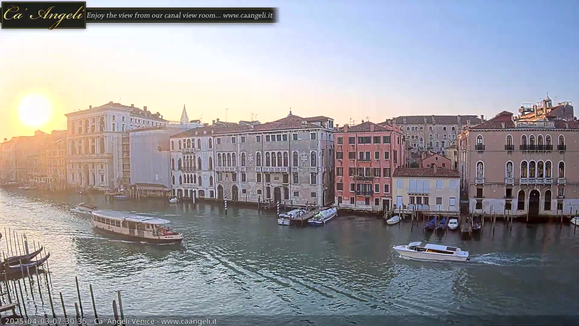 A sunrise illuminates a canal in Venice, with several boats on the water and numerous buildings lining the waterway.