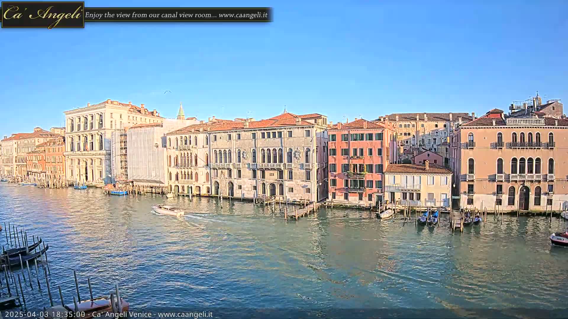 A sunny day in Venice shows a canal with colorful buildings lining its banks, several gondolas and a motorboat are visible on the water.