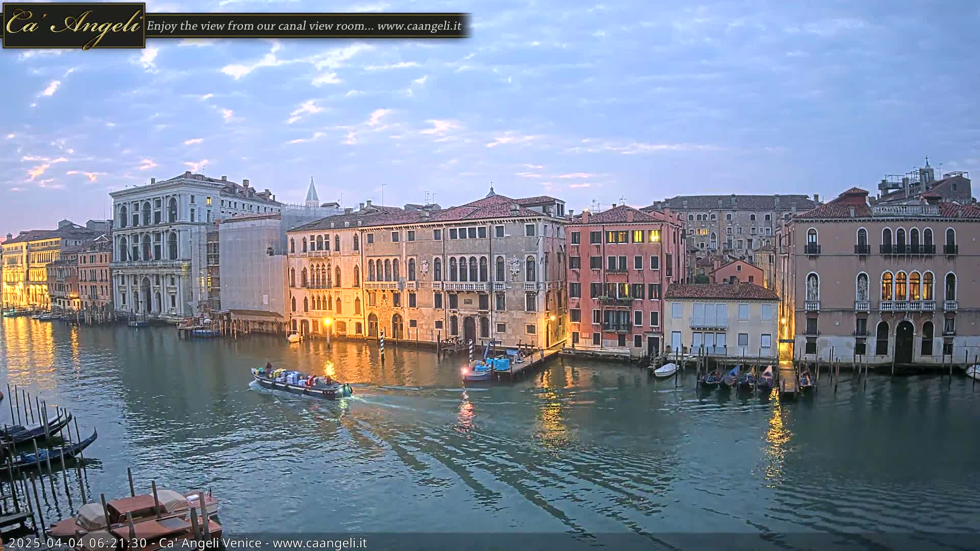 A tranquil canal scene in Venice at twilight, showing several buildings illuminated along the water's edge with boats moored and one small vessel underway.