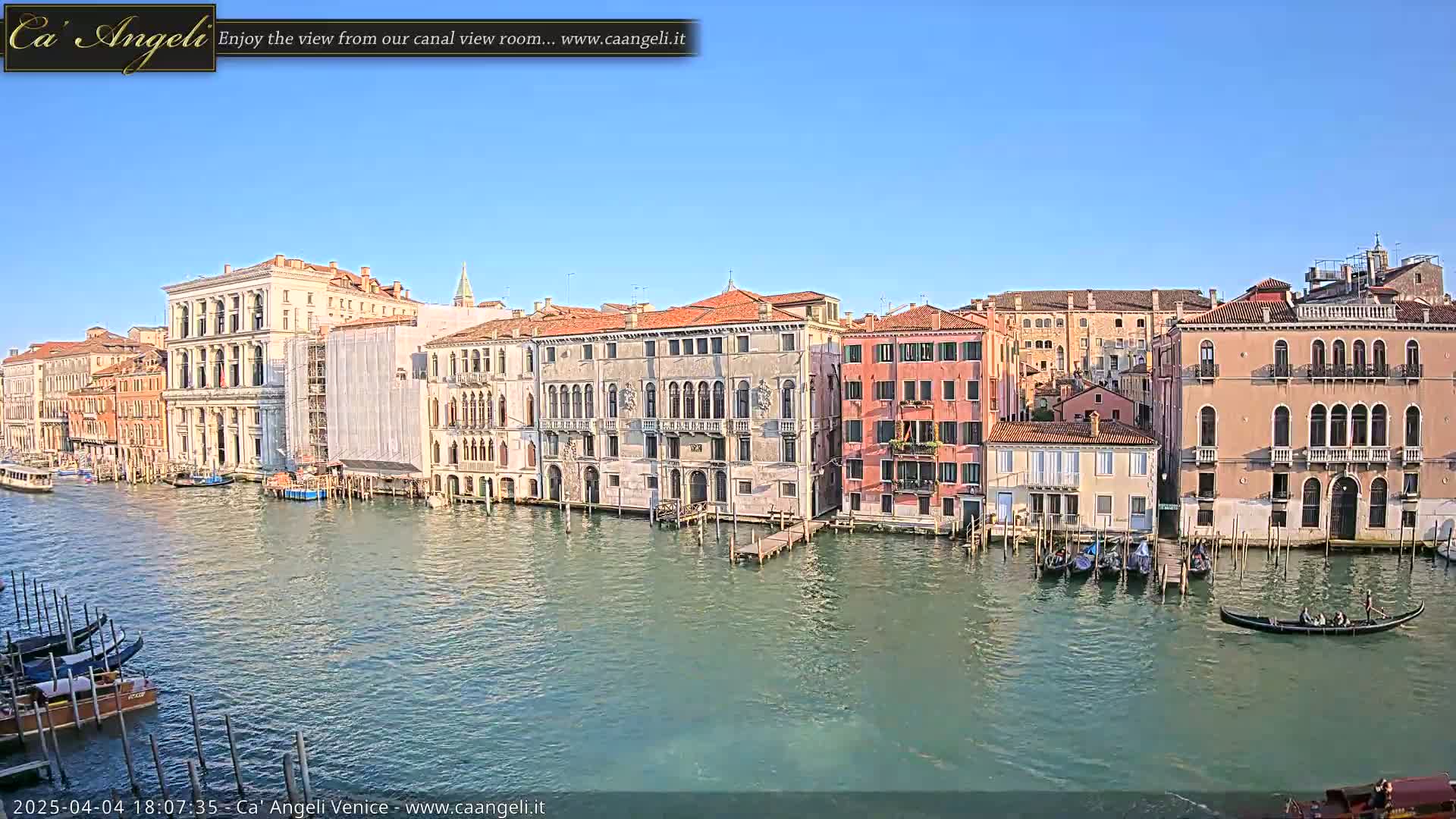 A sunny day in Venice shows a canal lined with several multi-story buildings, some with scaffolding, and several gondolas moored along the waterfront.