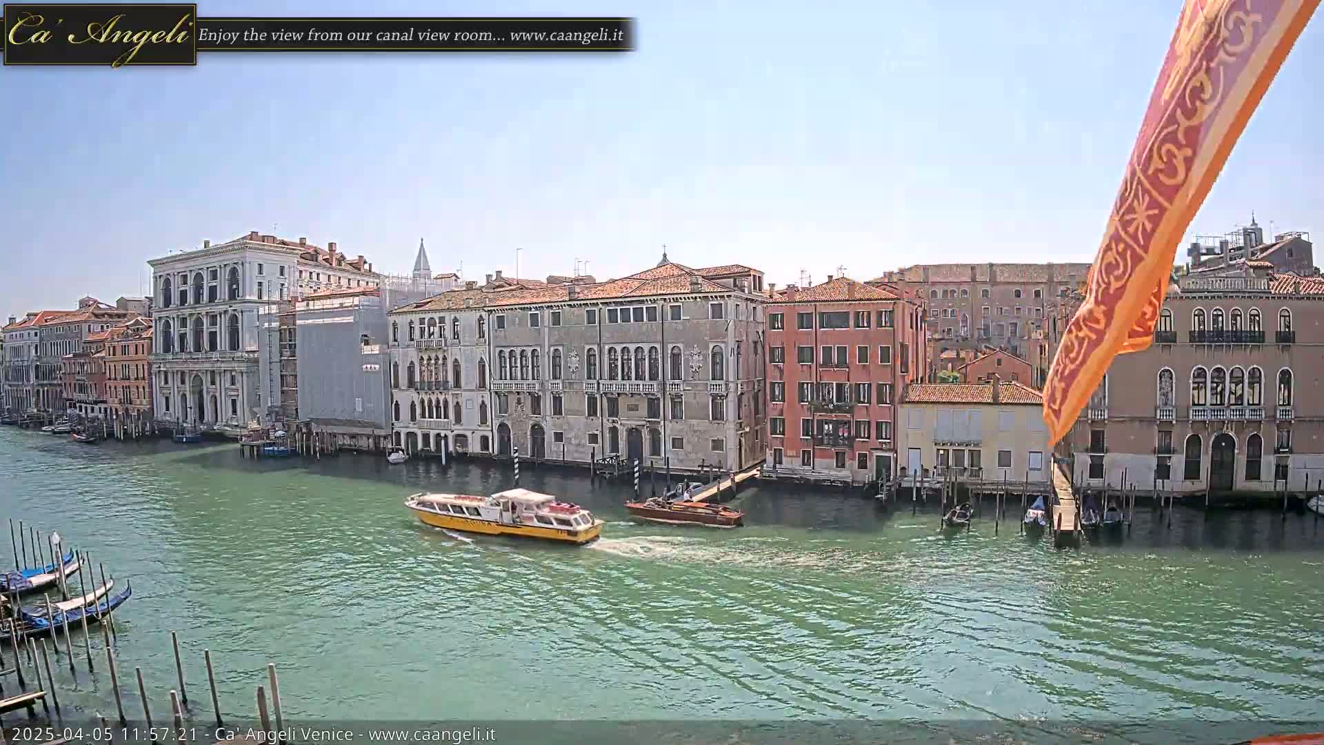 A sunny day in Venice shows a canal with several gondolas and motorboats passing by historic buildings.