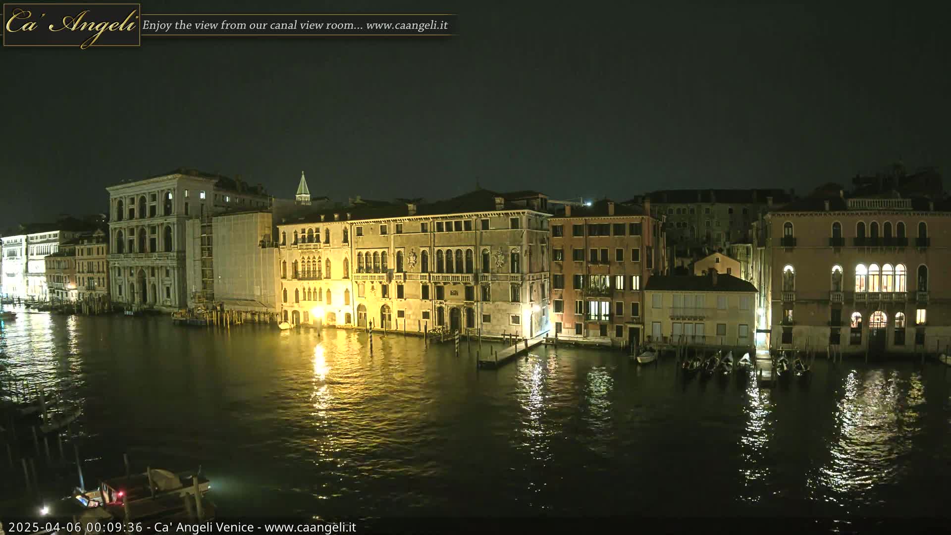 A nighttime view of a canal in Venice, Italy, shows several buildings illuminated along the waterway, with gondolas moored along the edge.