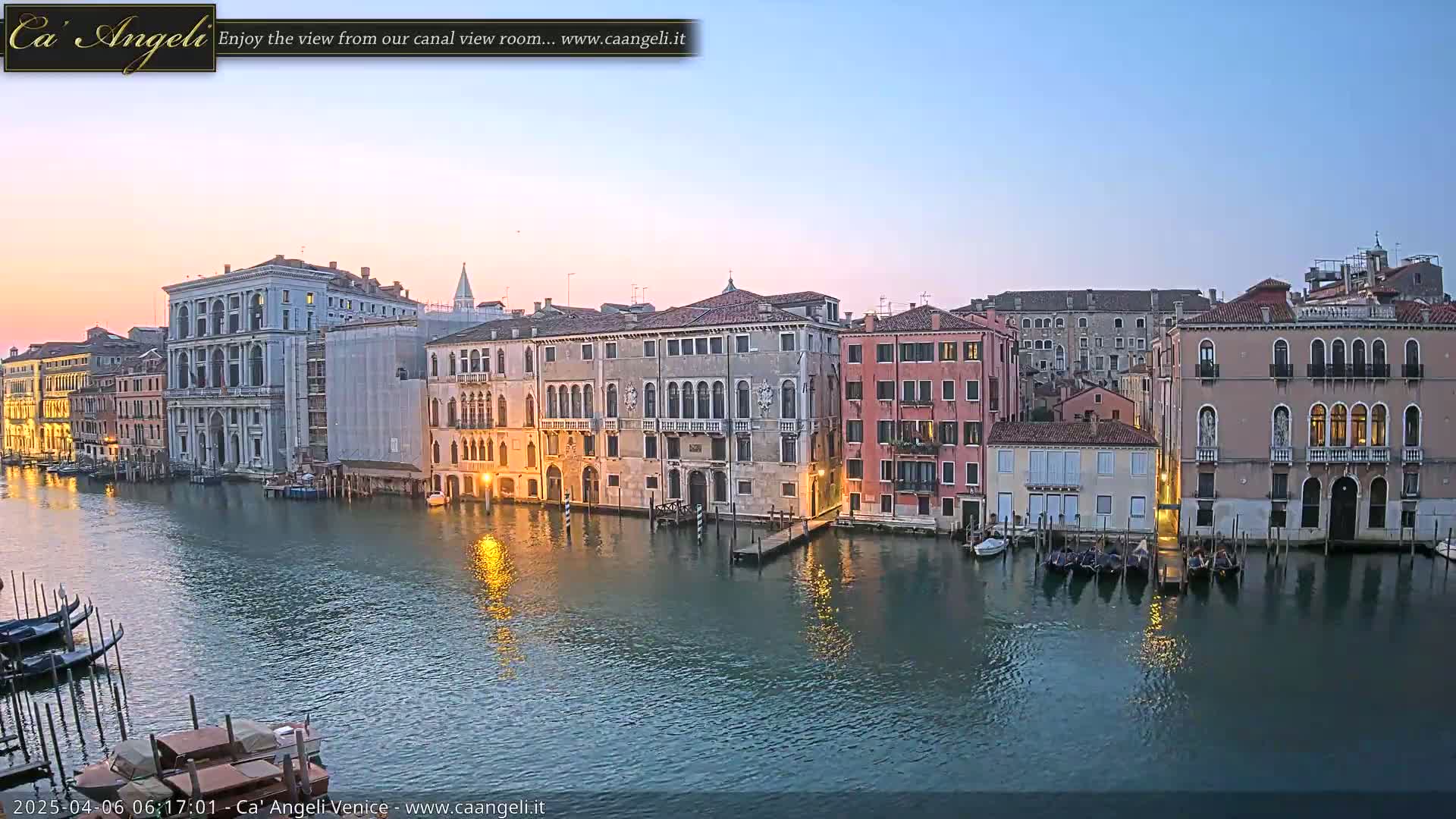 A calm canal in Venice at dawn is lined with historic buildings reflecting in the water, with several gondolas moored along the edge.